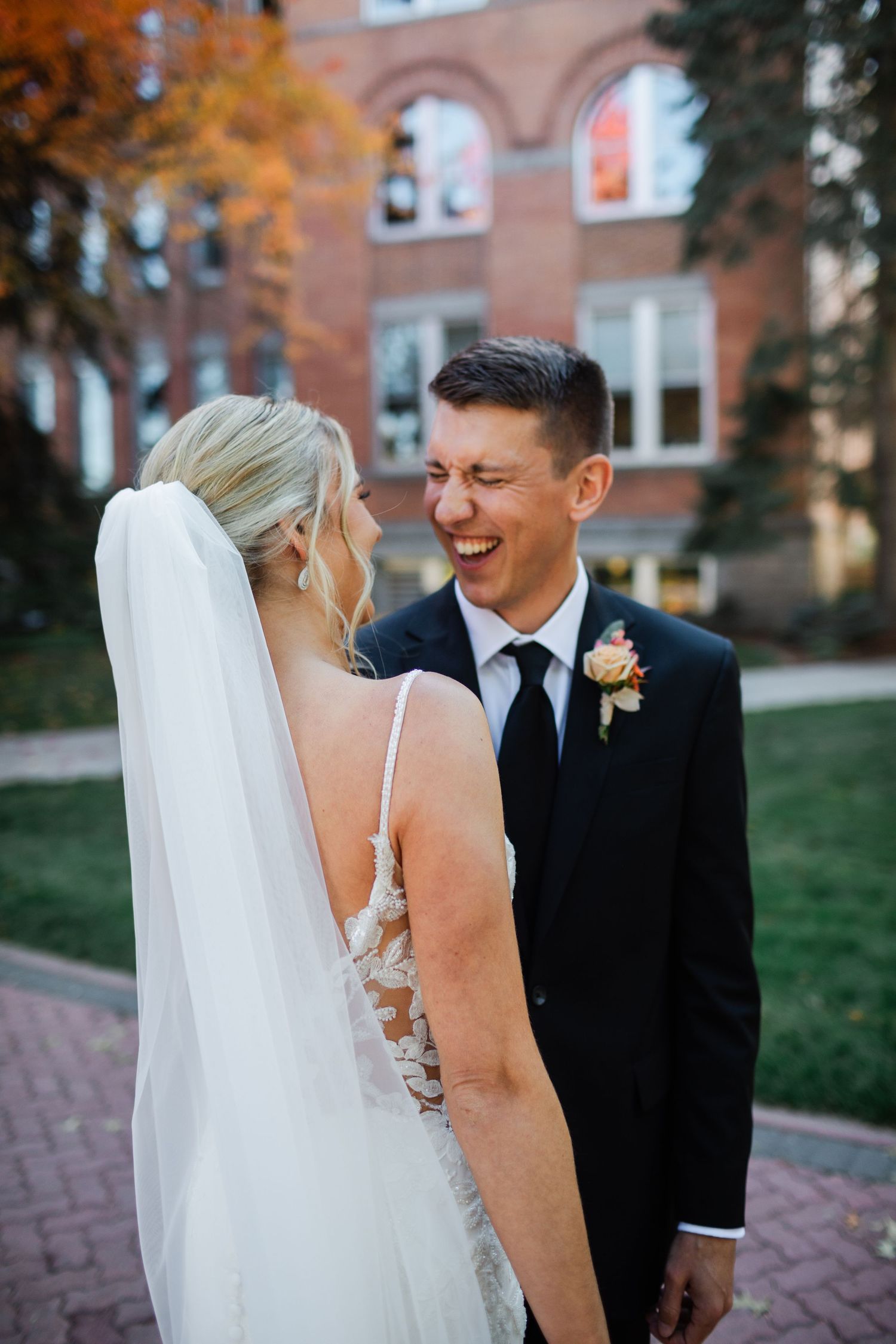Laughing groom in black suit with boutonniere faces bride in lace wedding dress and veil outside brick building.