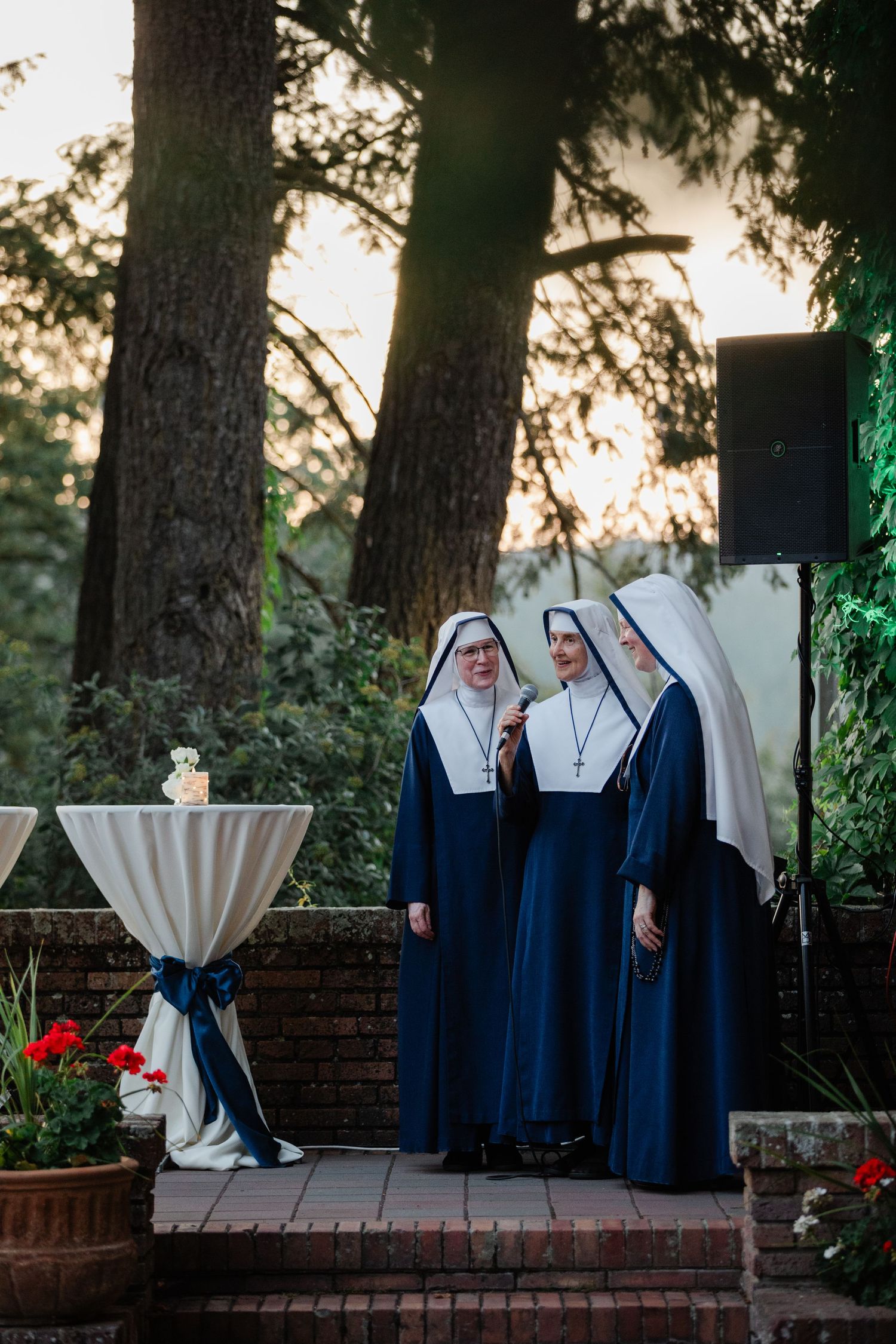 Three nuns in blue and white habits standing together outdoors at a garden event near a speaker.