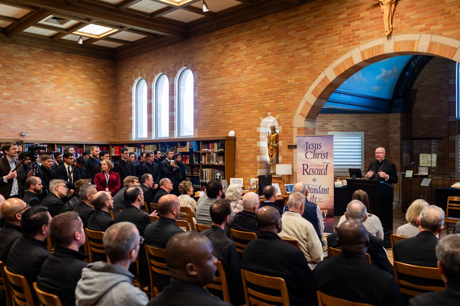 Clergy and attendees gather in a brick library hall beneath a Jesus Christ is Lord banner for a religious assembly.