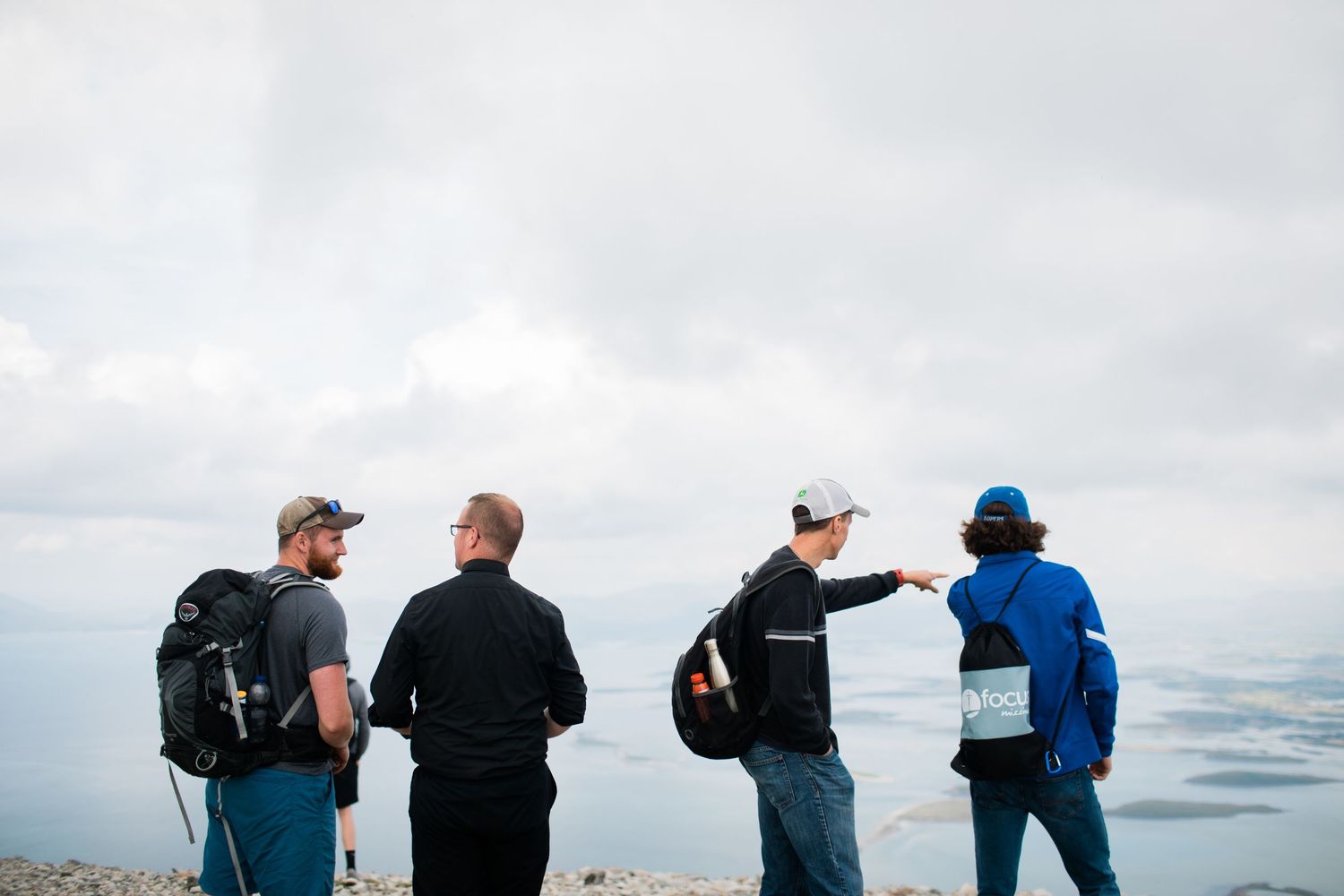 Four hikers stand on a mountain summit overlooking a vast landscape, one person pointing toward the horizon.