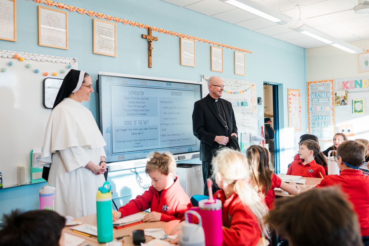 Catholic nun and bishop in black vestments visiting students in a colorful Catholic school classroom.