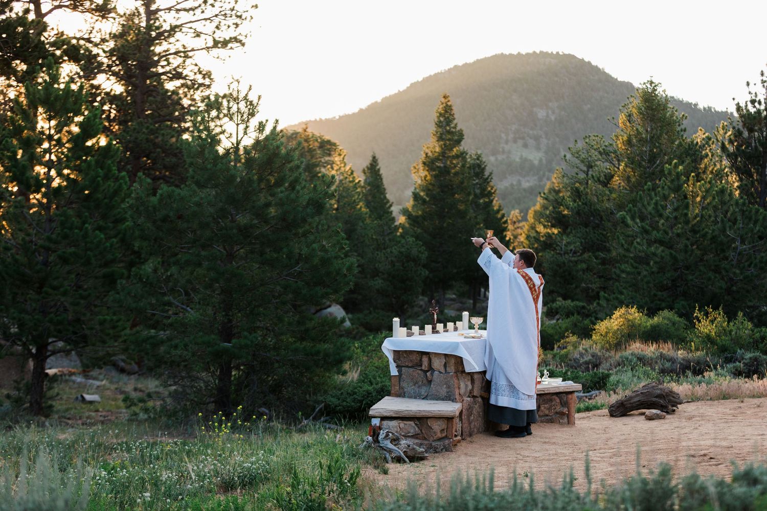 Priest celebrating outdoor Mass at stone altar surrounded by pine trees and mountain landscape at sunset.
