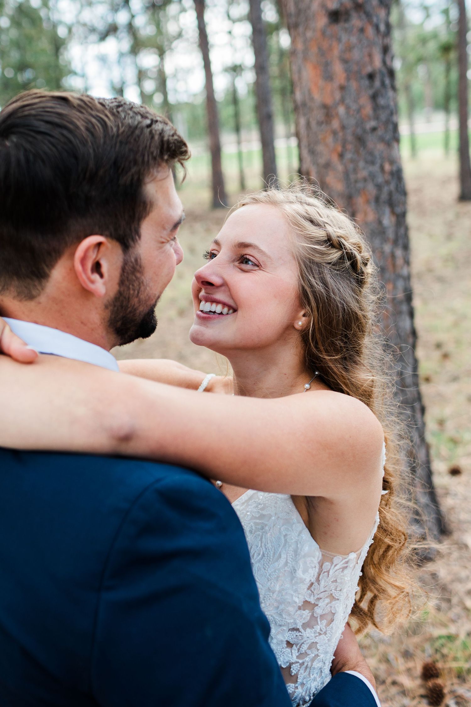 A smiling bride in a lace dress gazes lovingly at her groom in a navy suit during an outdoor forest wedding.