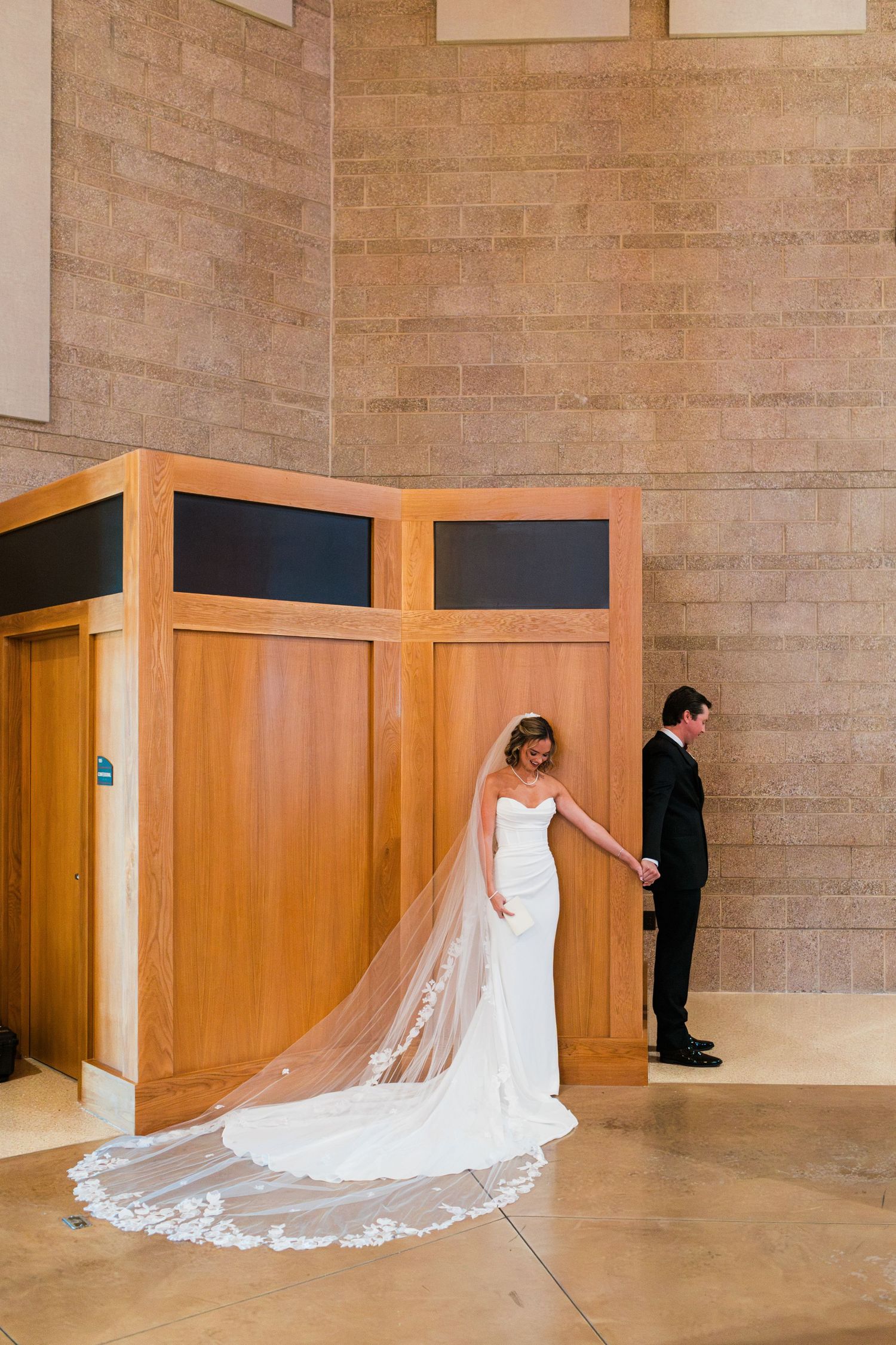 Bride in a white gown with cathedral veil holds grooms hand during first look near wooden partition.