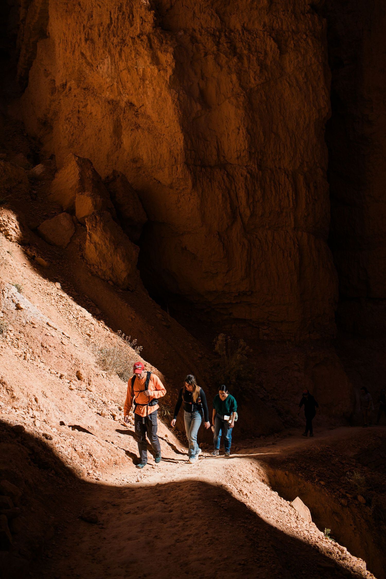Hikers traverse a sunlit path through a dark desert canyon, casting dramatic shadows on the rocky terrain.