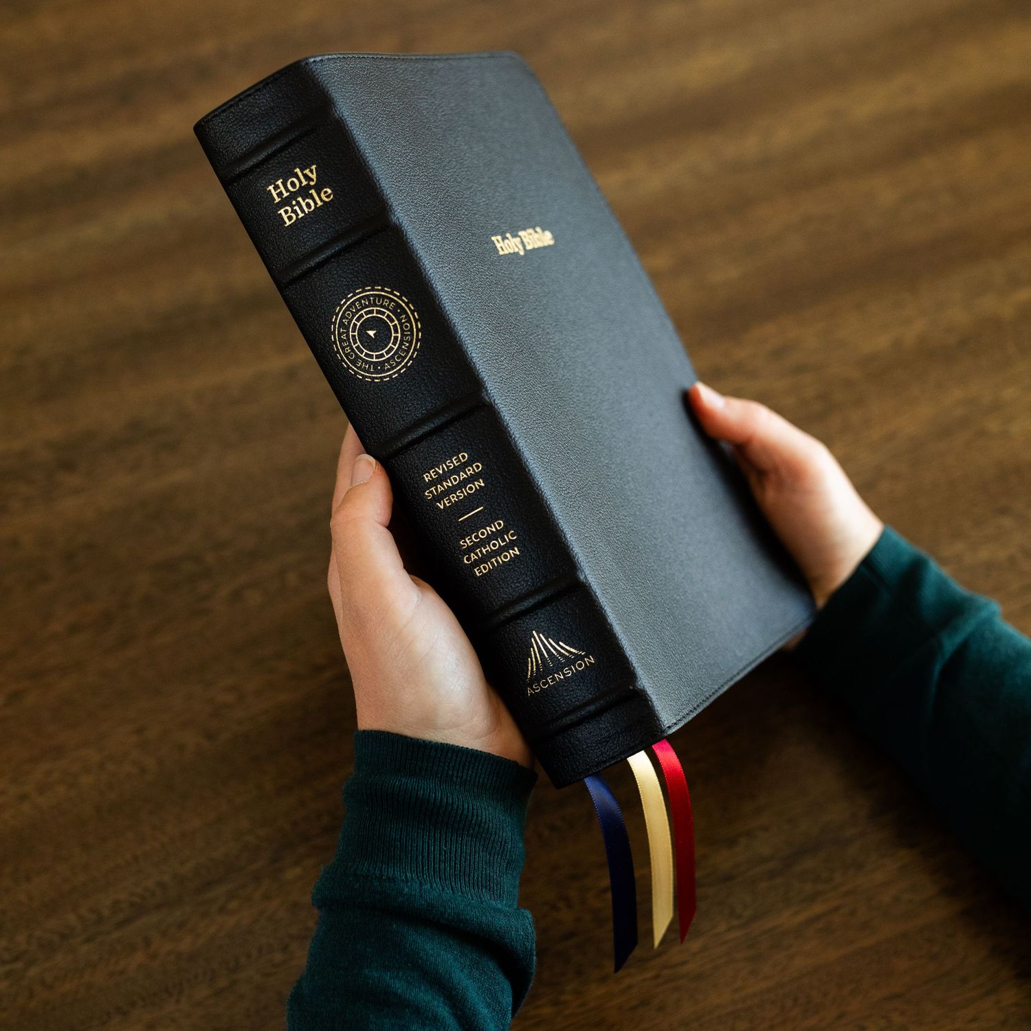 A hand holds a black leather Bible with gold edges and a bookmark ribbon against a wooden table surface.