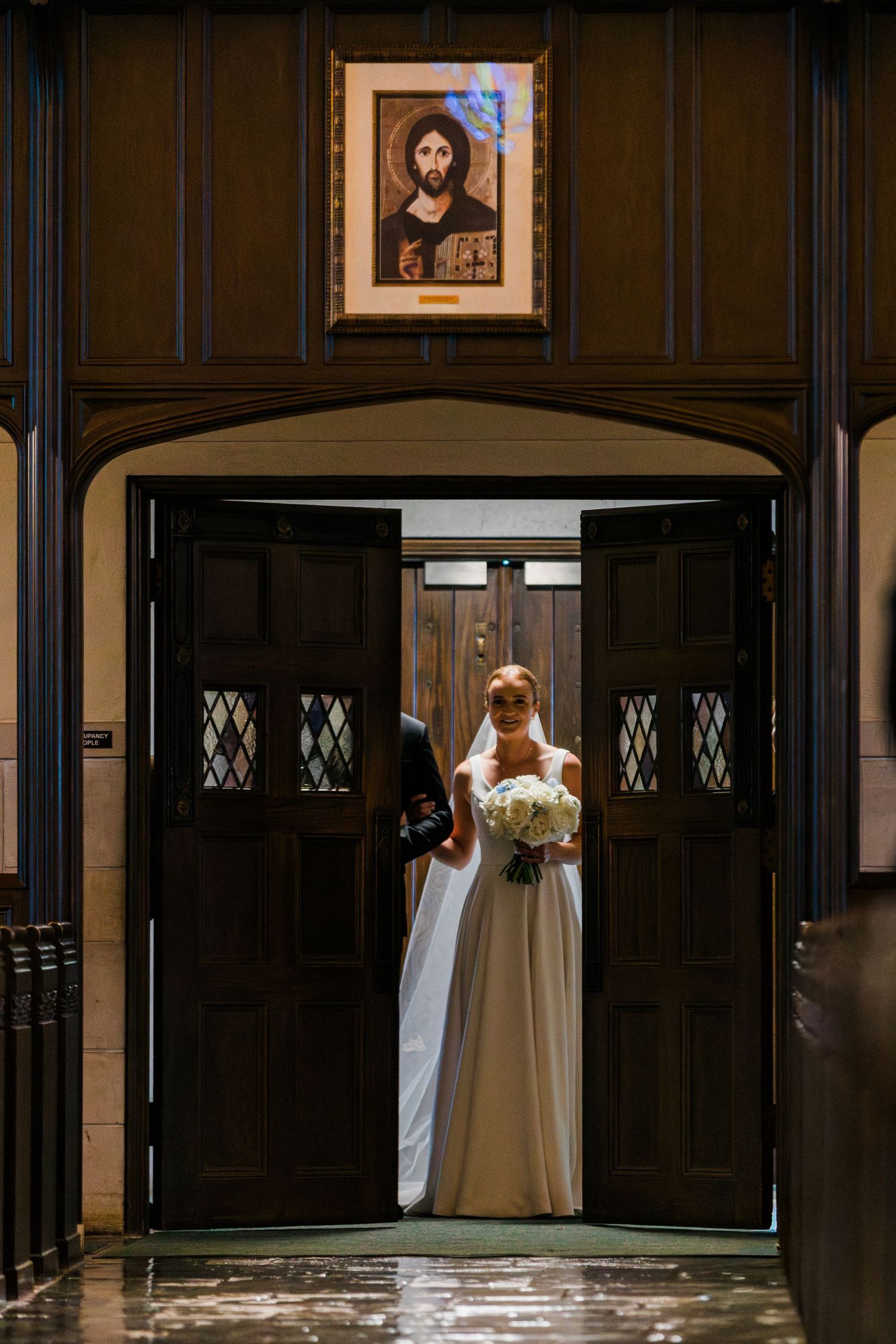 Bride in white gown holding bouquet stands at ornate church doors beneath religious icon painting.