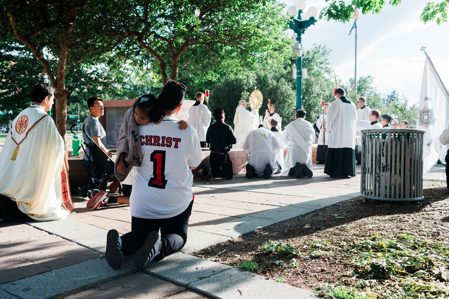 Person kneeling in a Christ jersey at an outdoor Catholic Mass ceremony with priests in white vestments.