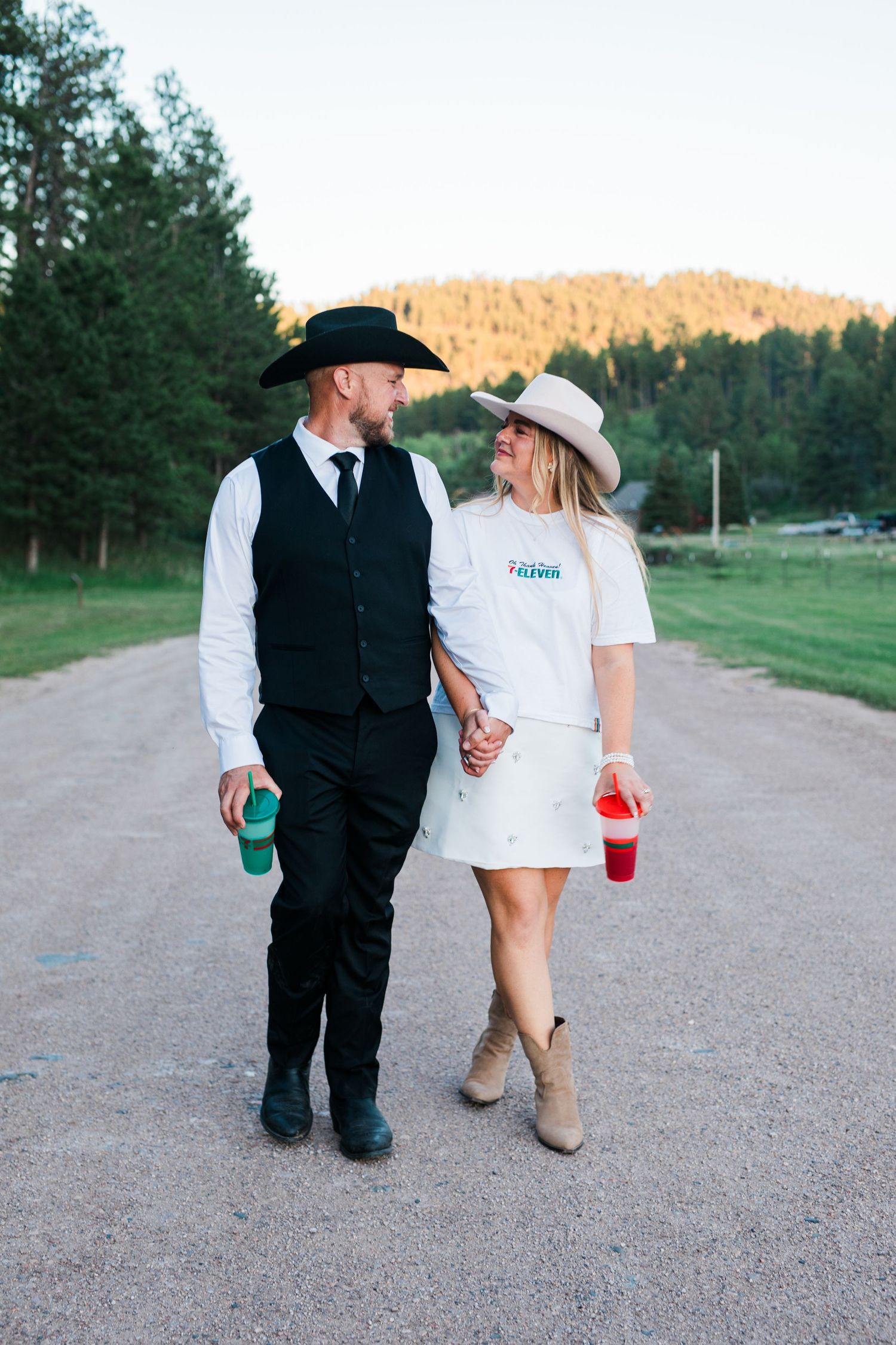 Couple in cowboy hats walking hand-in-hand down a rural dirt road surrounded by pine trees and mountains.