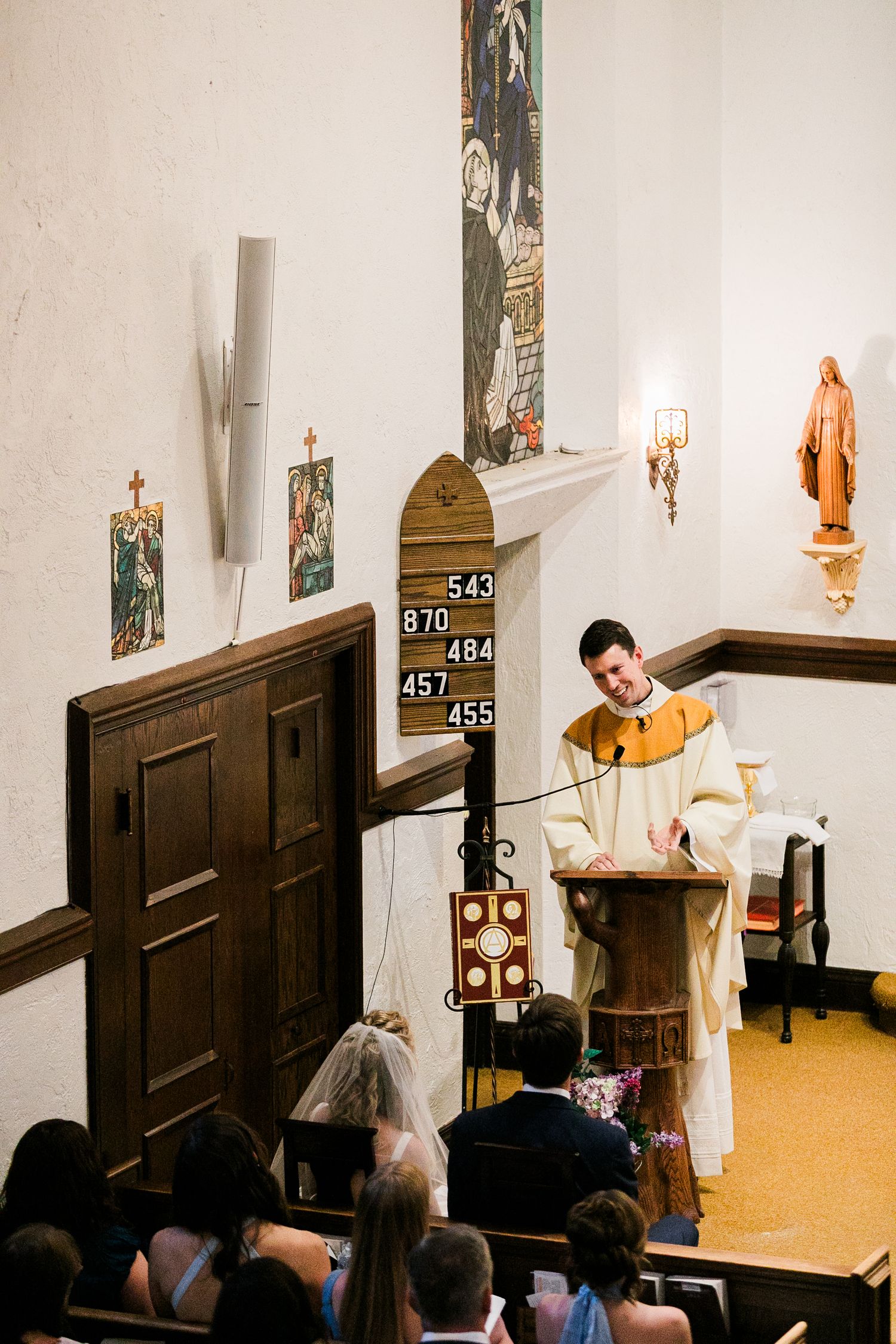 Priest officiates wedding ceremony at wooden pulpit in white chapel with hymn board and religious artwork.