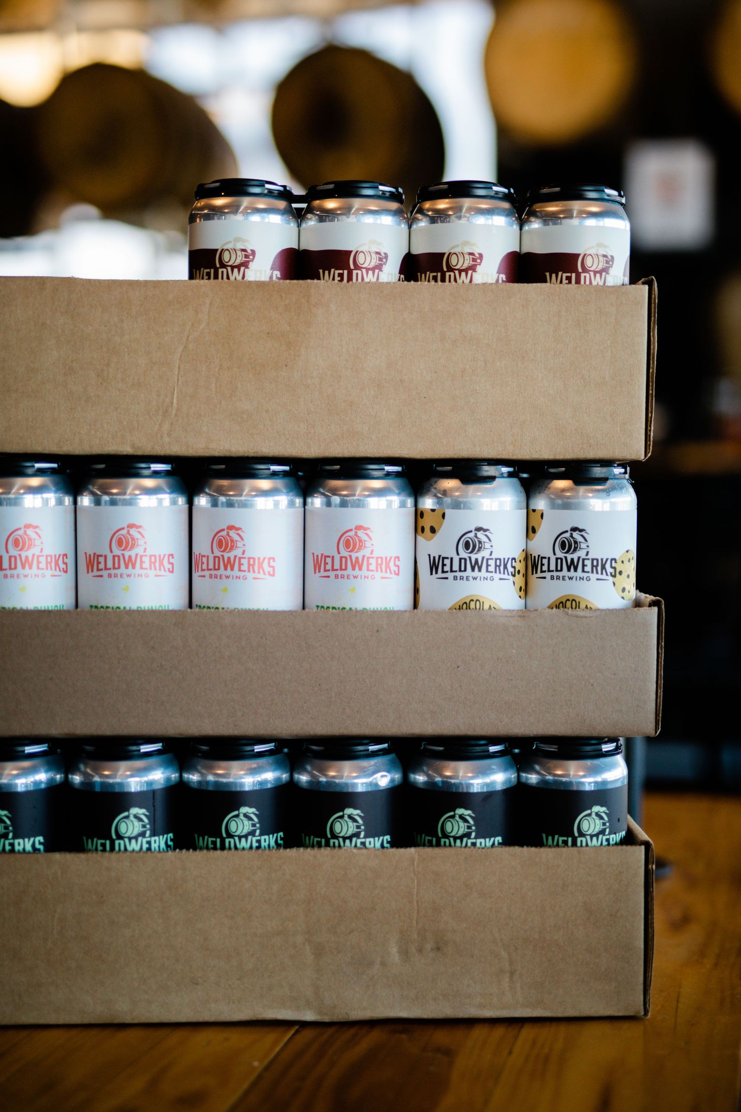 Wooden shelves display rows of craft beer cans in a rustic brewery setting with soft bokeh lighting in background.