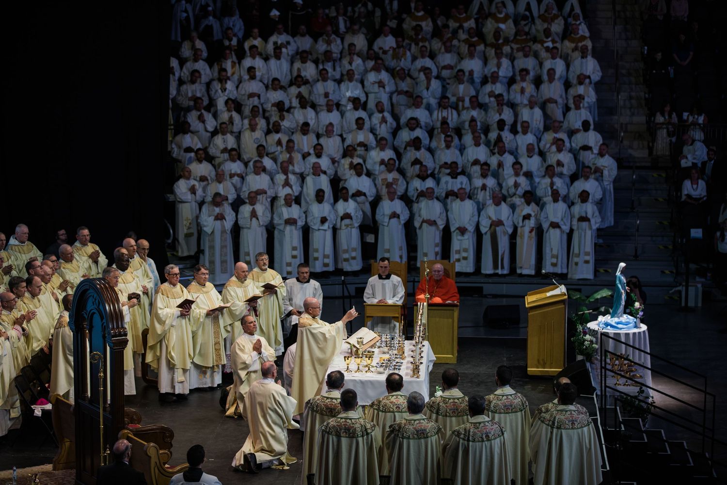 Grand Catholic Mass ceremony with clergy in white vestments performing at a large indoor arena.