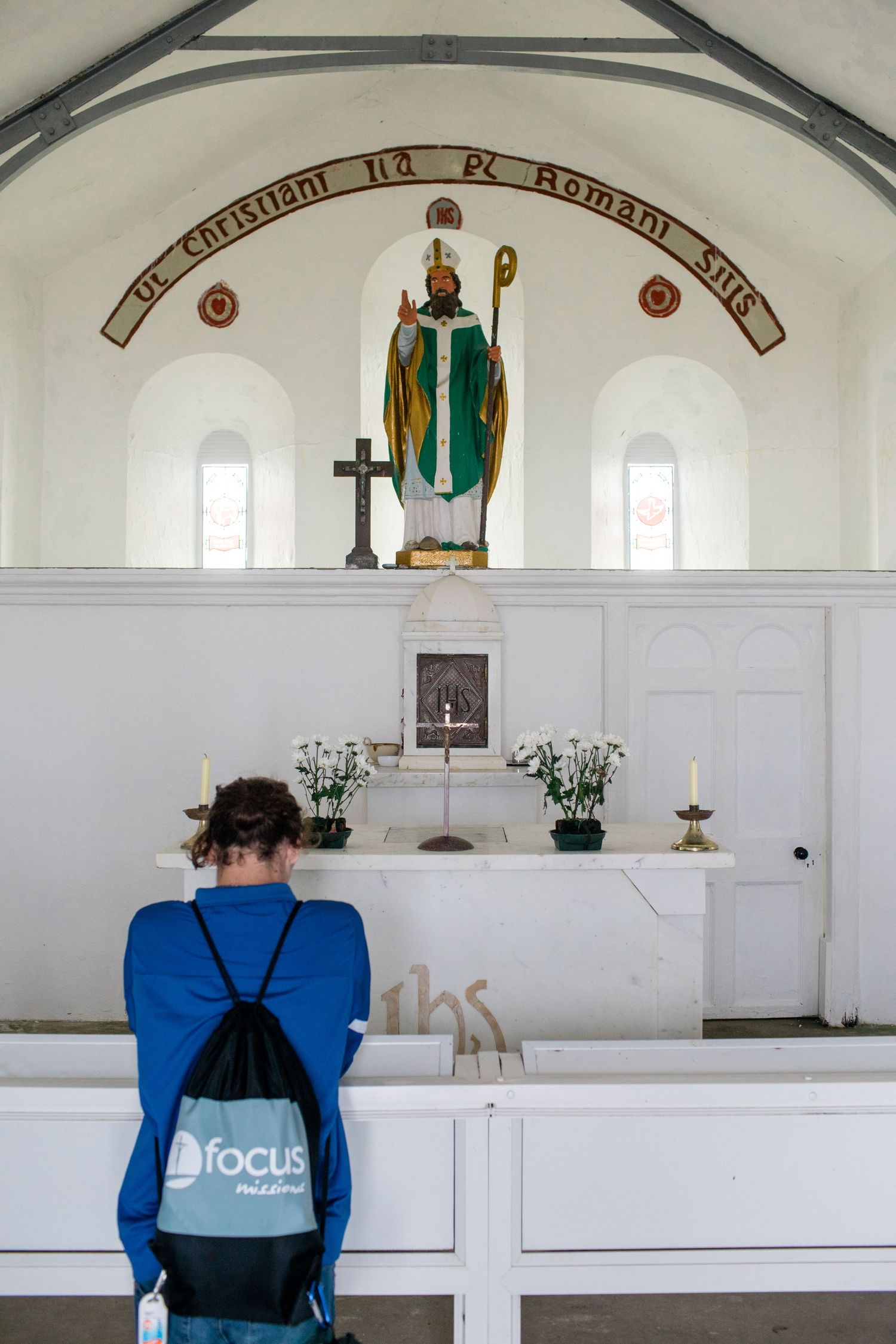A person praying before a white altar with a Saint Patrick statue in a small chapel.