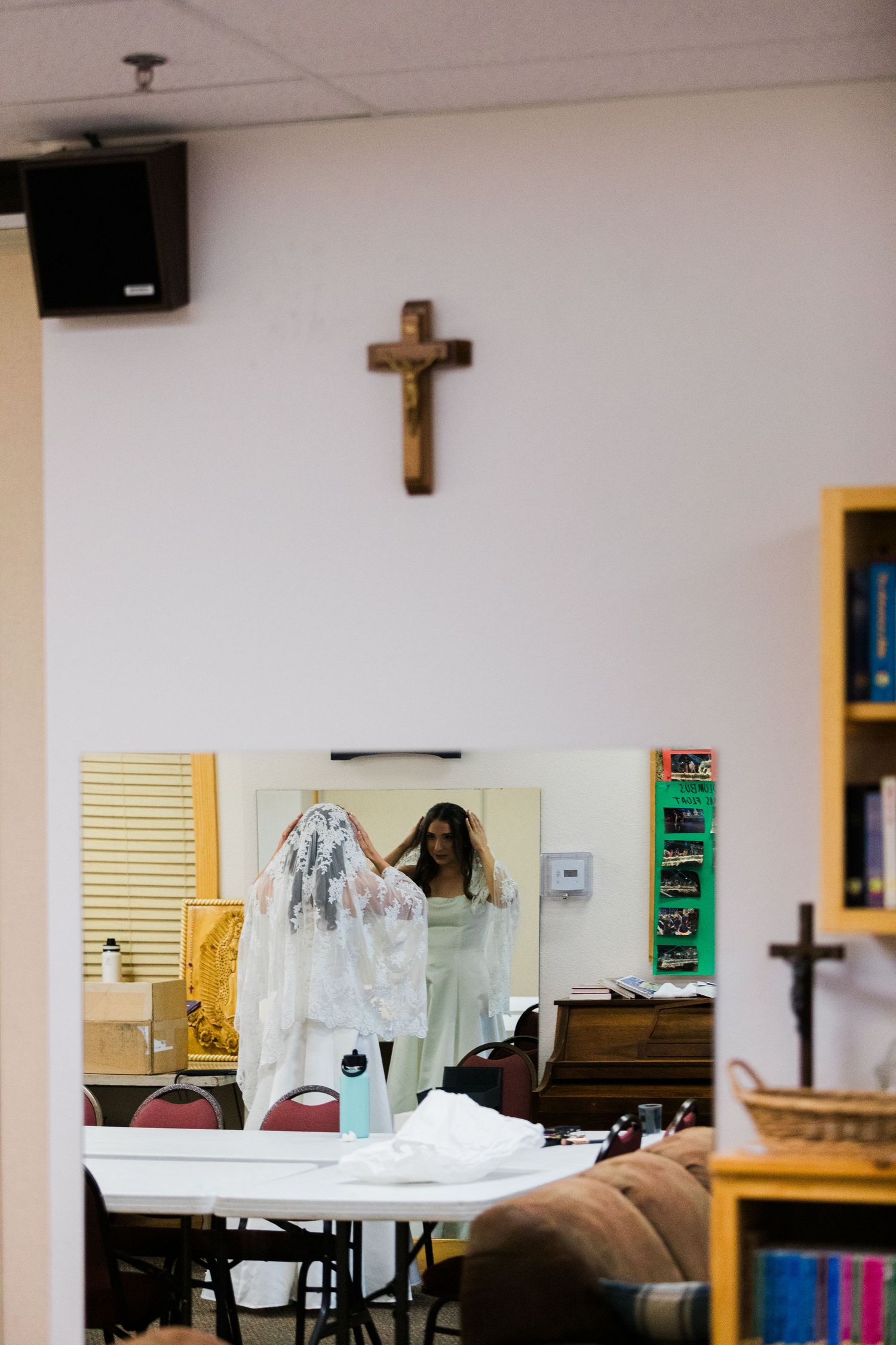 Woman holding a wedding veil in front of a mirror inside a church room with a wooden crucifix on the wall.