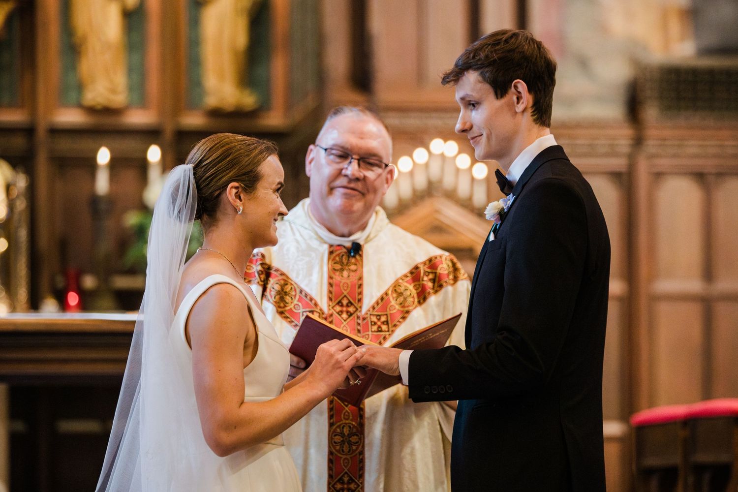 Bride and groom exchange rings during a church wedding ceremony with a smiling officiant in ornate robes.