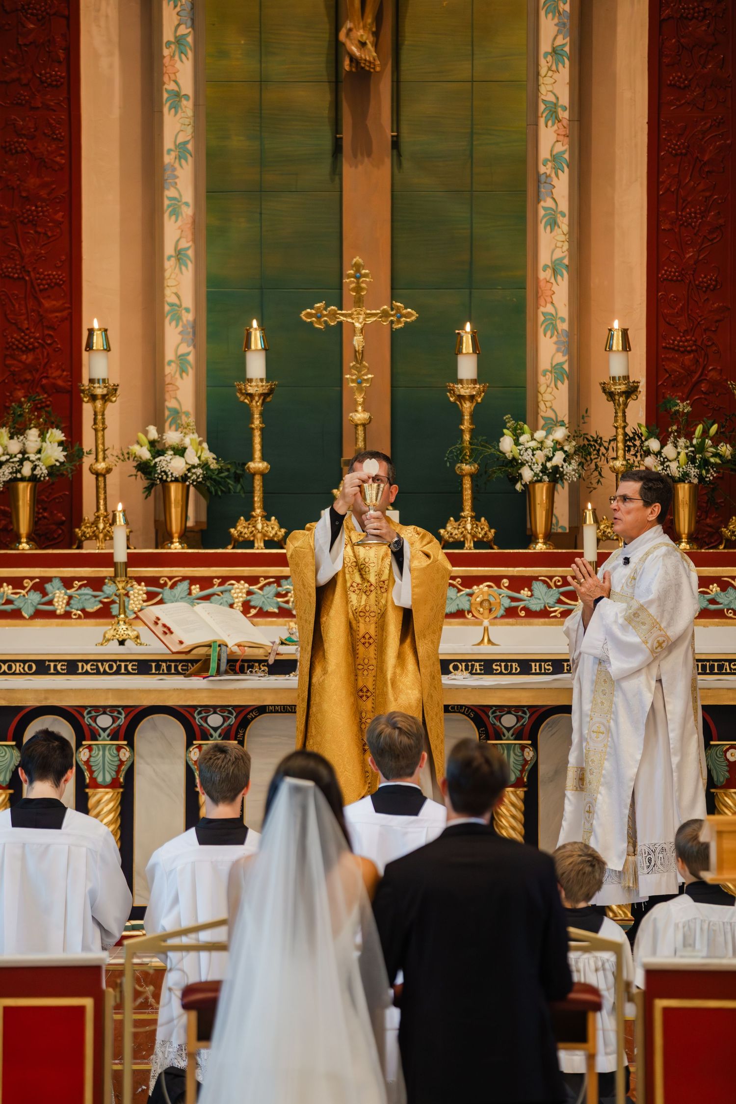 Clergy members in white and gold robes conduct a traditional church ceremony at an ornate altar with candles and flowers.
