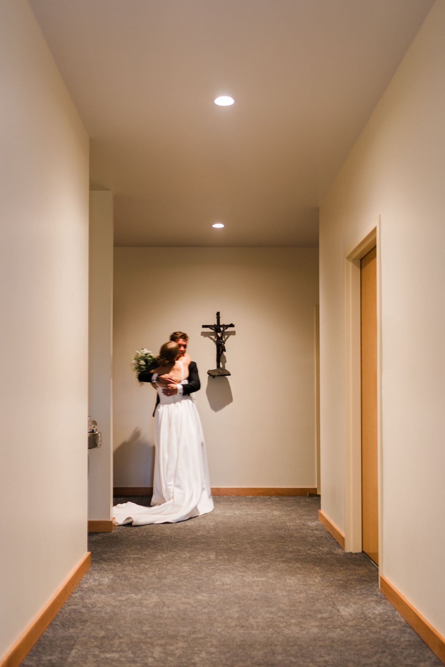A hallway with beige walls, recessed lighting, and a crucifix mounted on the wall.
