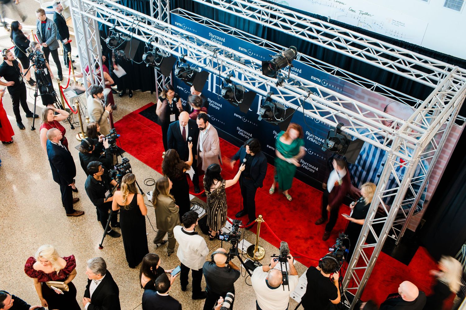 Overhead view of a formal event with people mingling on a red carpet under white truss lighting structures.