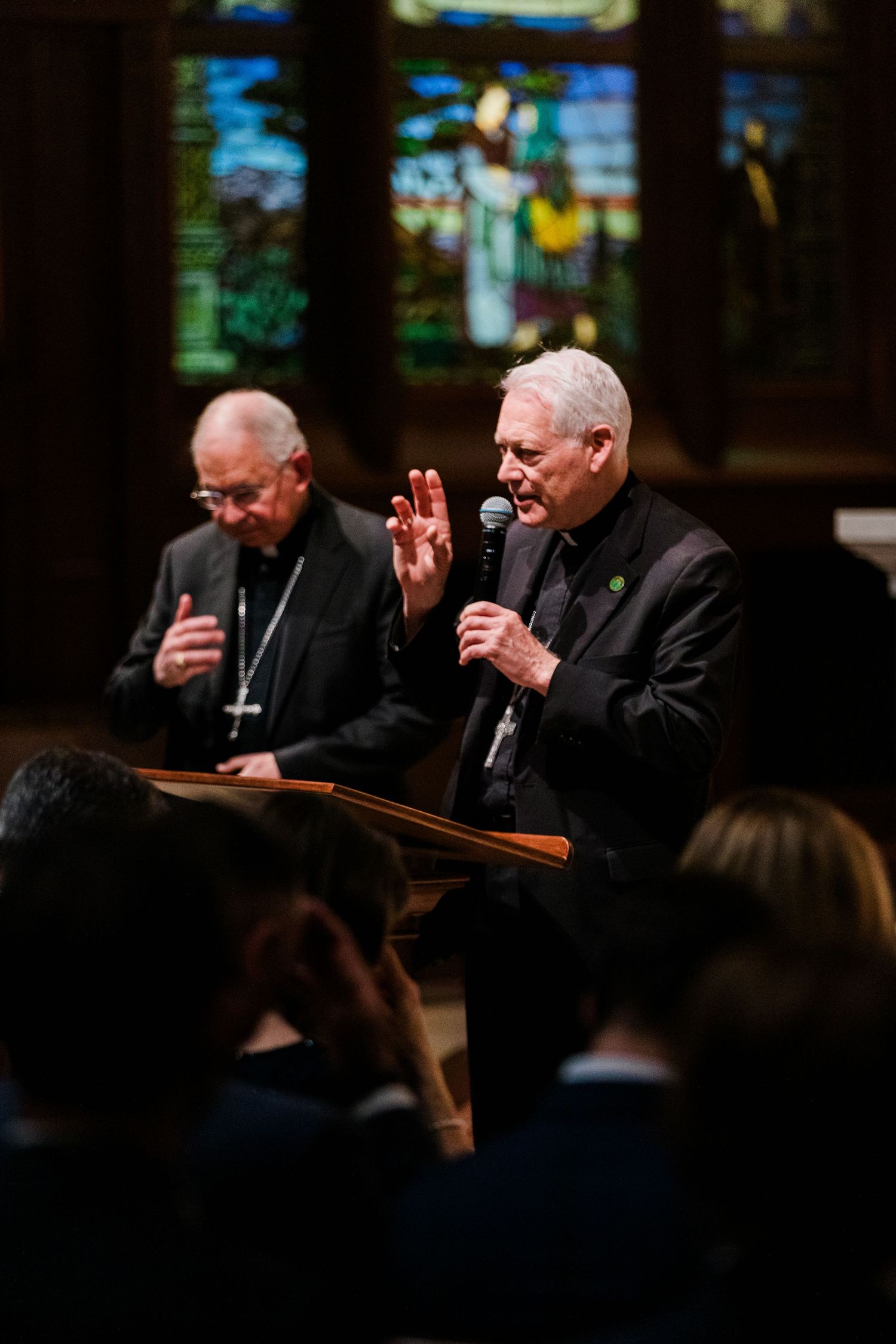 Two clergy members in black robes speaking at a podium during a religious service in a church with stained glass windows.