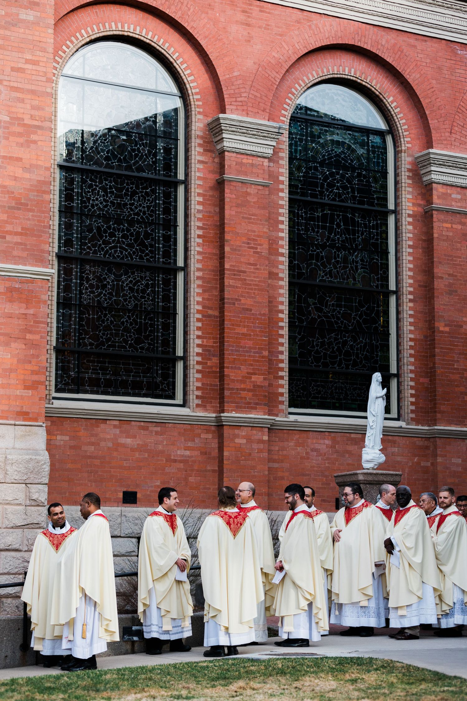 Row of people wearing cream-colored ceremonial robes stand in line against a red brick church exterior.