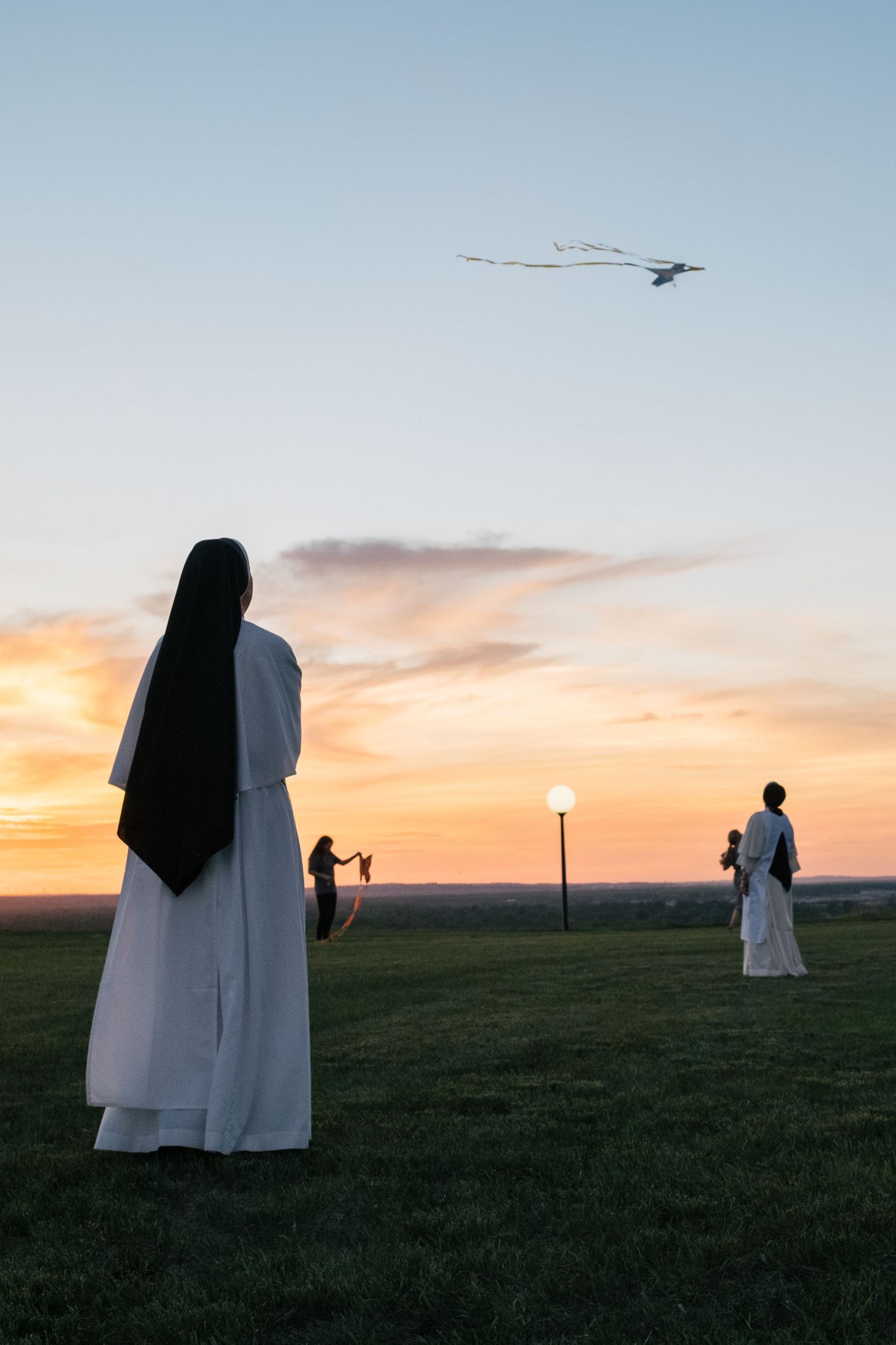 Silhouettes of nuns stand in a field against a sunset sky with an airplane flying overhead.