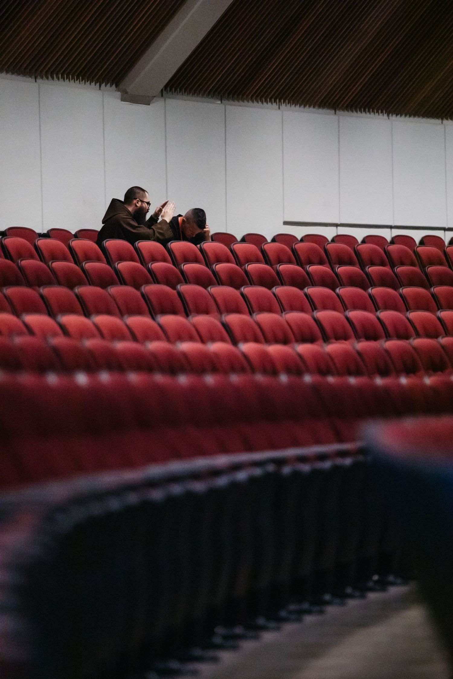 Empty red theater seats with figures visible in distant upper rows.