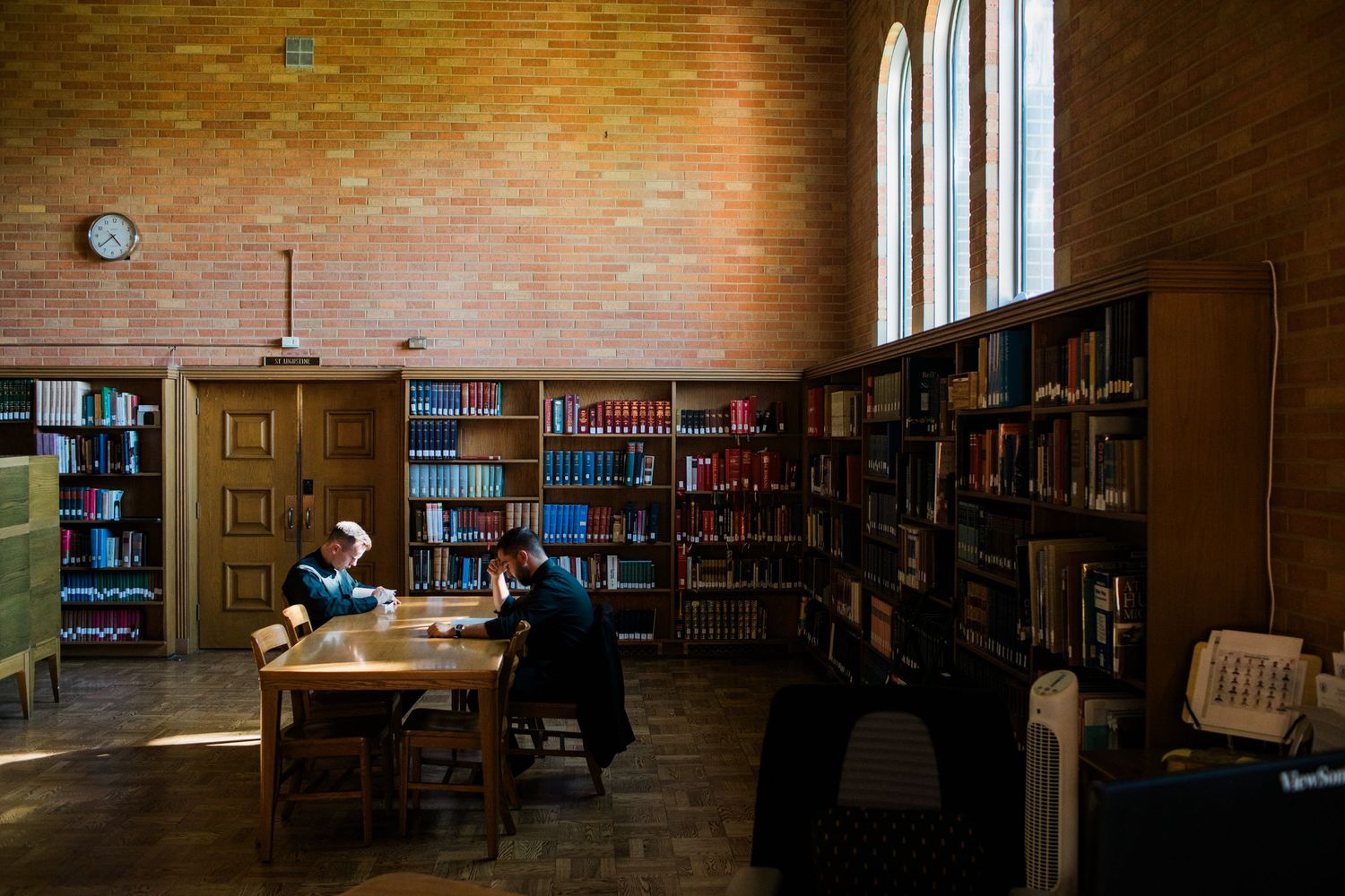Students study among tall bookshelves in a warmly lit brick-walled library with arched windows and a wall clock.