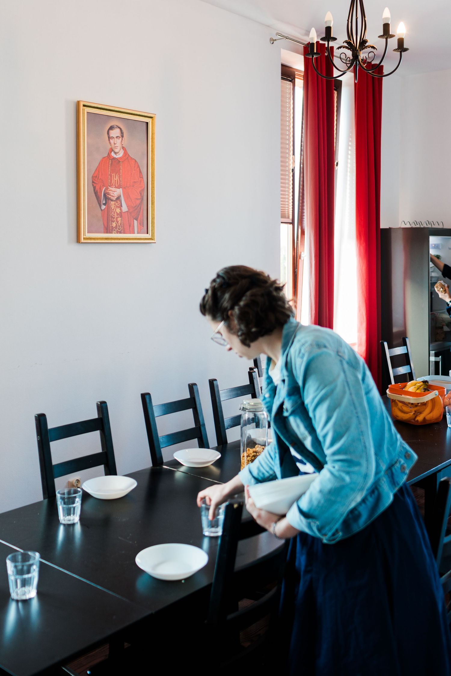 Woman setting a dining table beneath a framed Catholic saint portrait with red curtains in background.