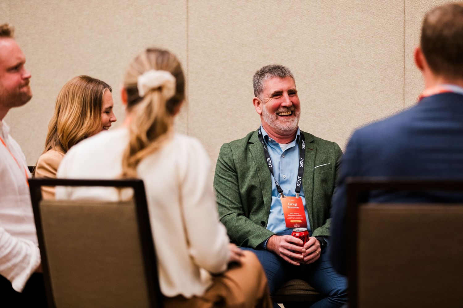 A man in a green blazer laughs during a conference networking session with colleagues seated in chairs.