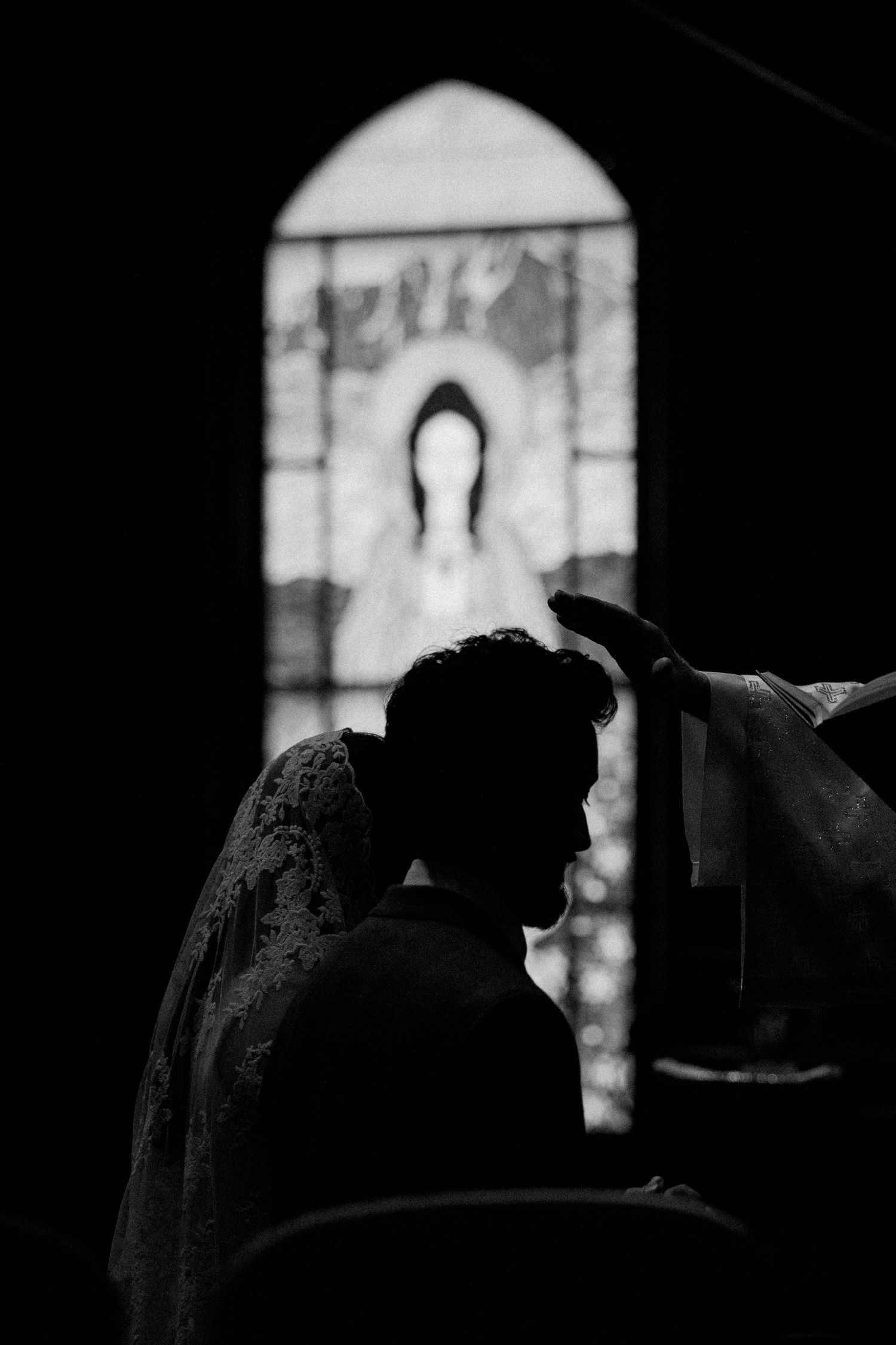 Black and white photo of a wedding ceremony in a church with stained glass window featuring Virgin Mary silhouette.