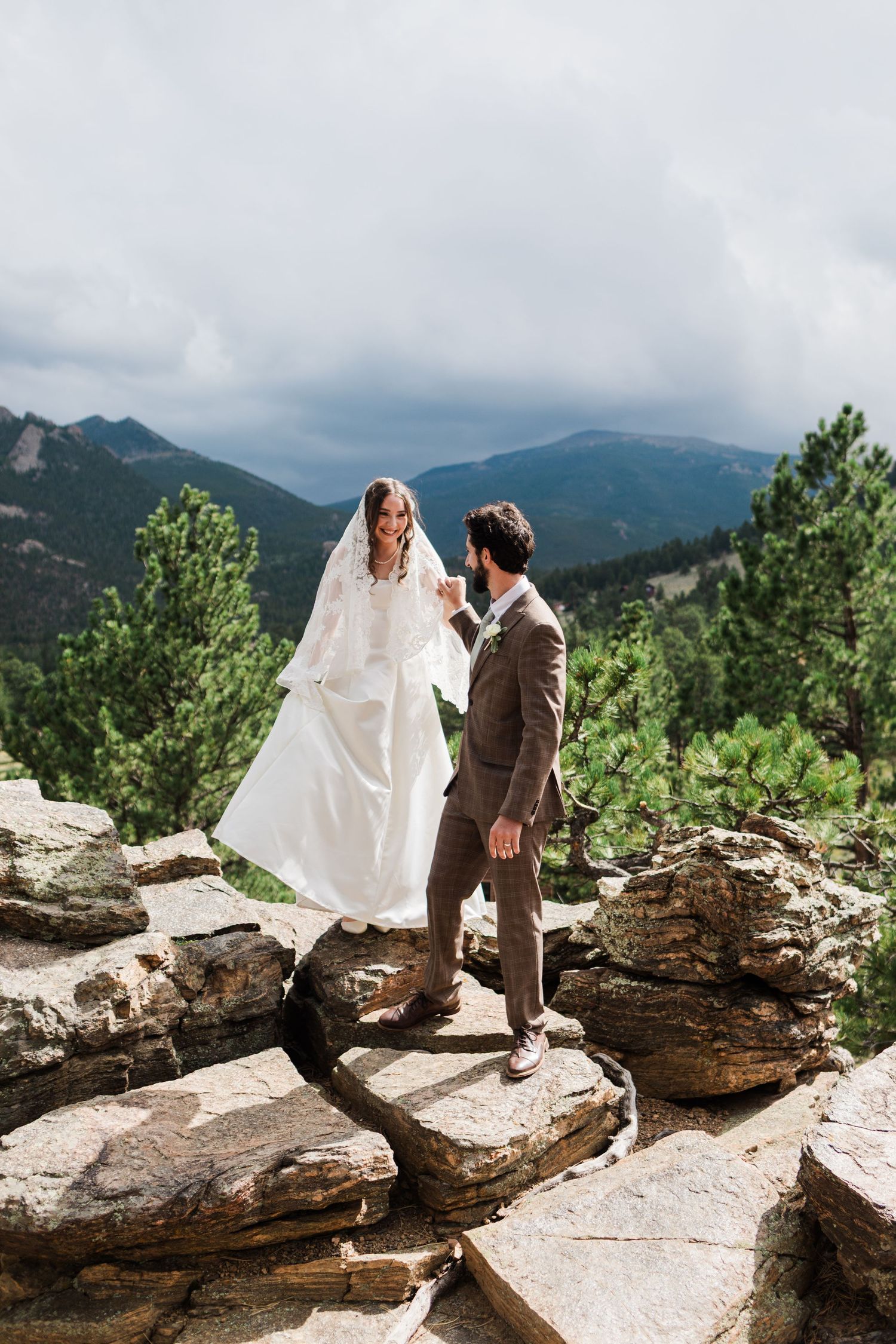 Bride and groom stand on rocky mountain overlook, bride in white gown and veil, groom in brown suit, evergreen trees behind.