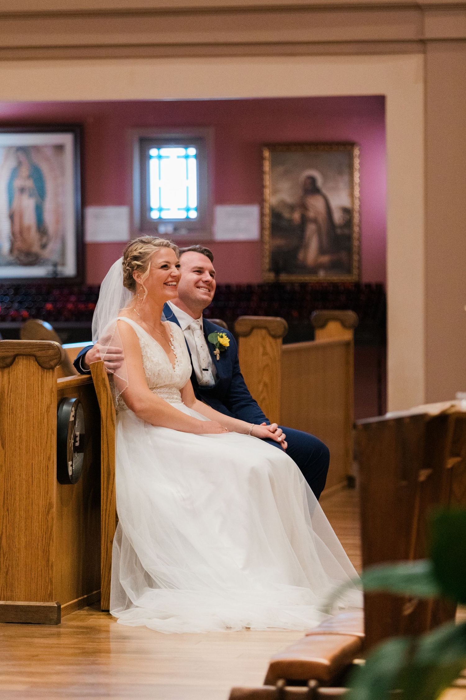 Bride and groom sitting together in church pew, smiling joyfully during wedding ceremony with religious artwork in background.