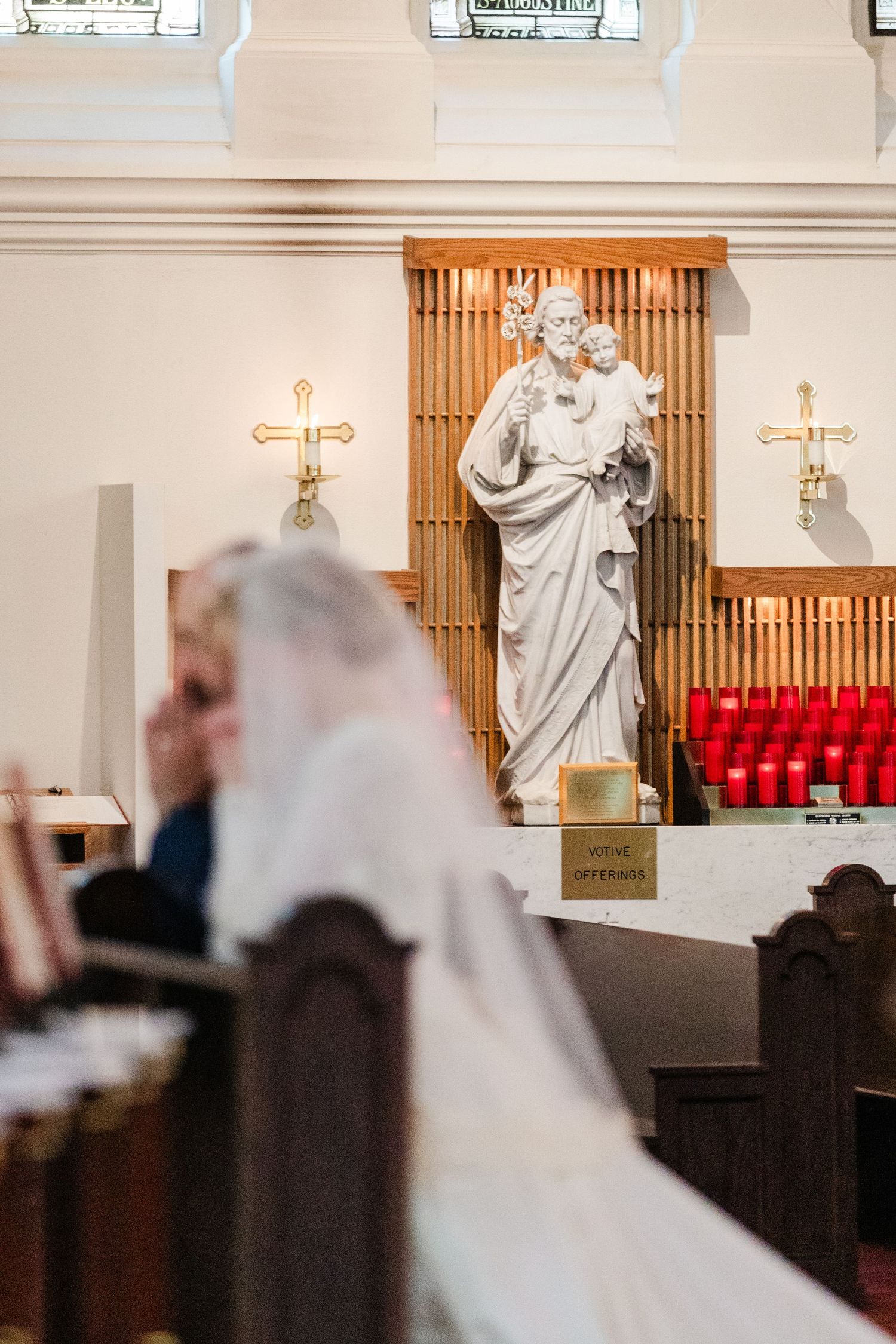 Bride in white veil seated in church pew with Saint Joseph statue and red candles on altar in background.