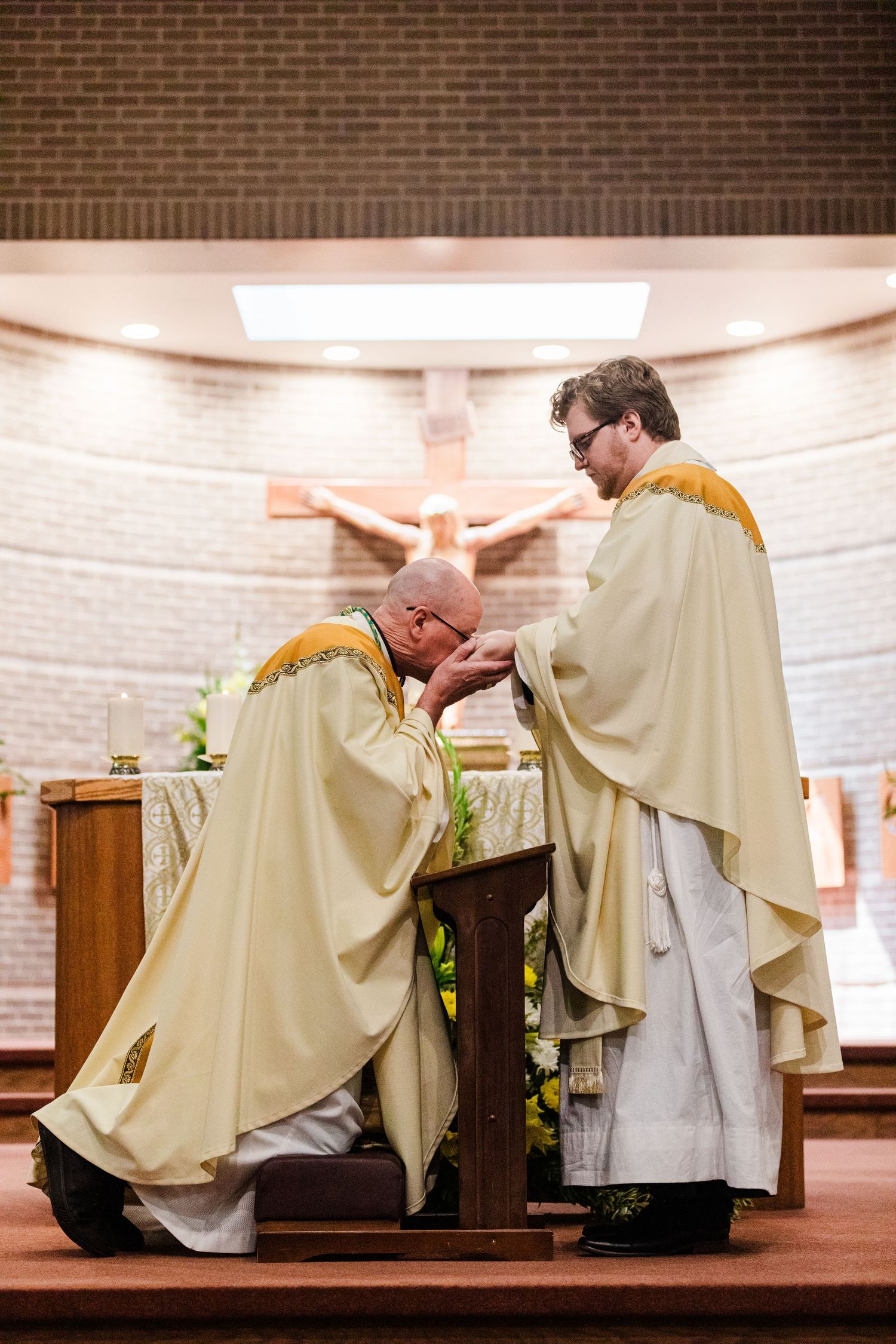 Two Catholic priests in cream vestments perform a religious ceremony at the altar with a crucifix in the background.