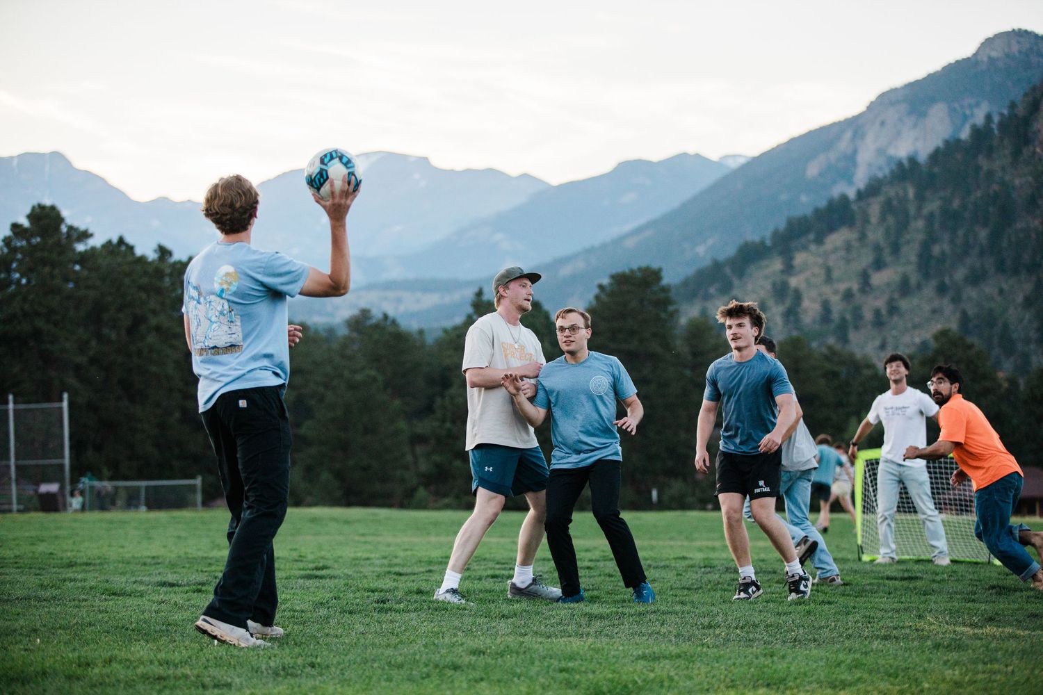 Group of young adults playing handball on a green field with scenic mountain backdrop at dusk.