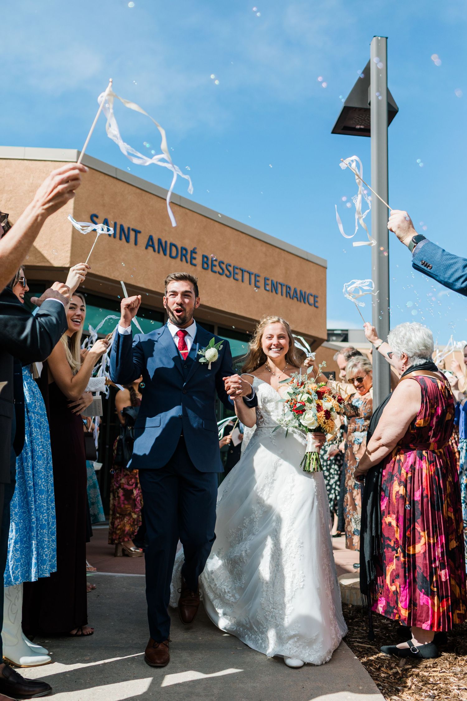 Newlyweds exit Saint André Bessette church amid cheering guests waving ribbon wands and bubbles on a sunny wedding day.