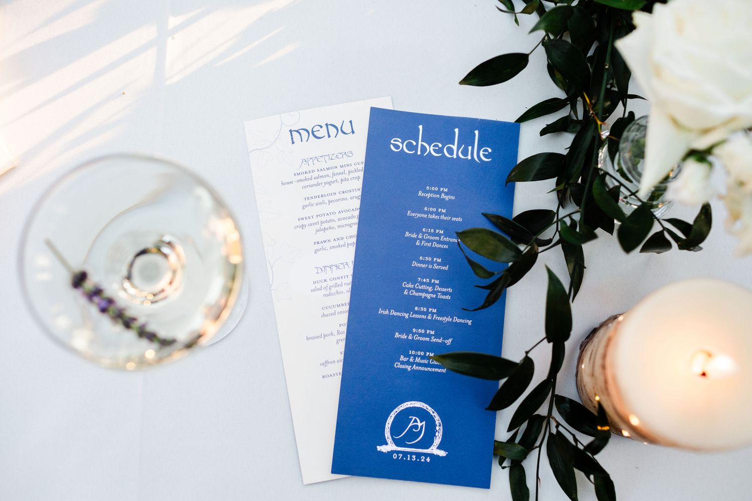 Elegant wedding menu and schedule cards displayed beside a wine glass, candle, and white floral arrangement.