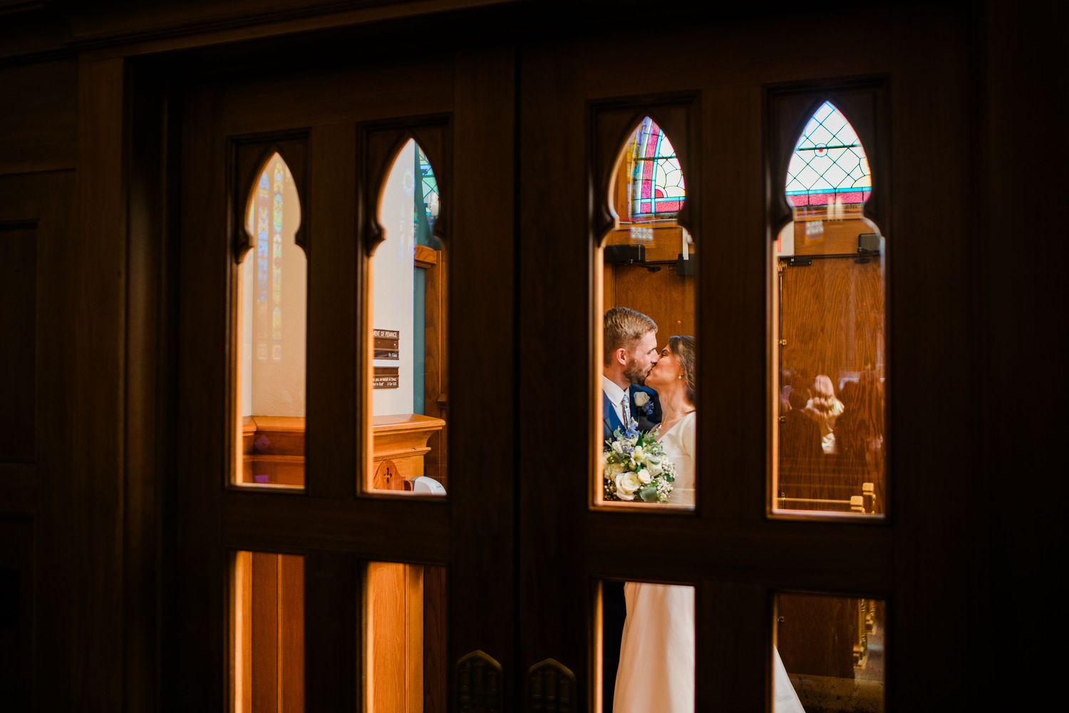 Couple sharing a kiss viewed through ornate church doors with stained glass windows during wedding ceremony.