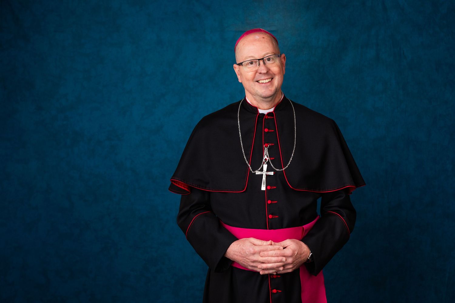 Catholic bishop in black cassock and pink zucchetto smiling warmly against a blue background.