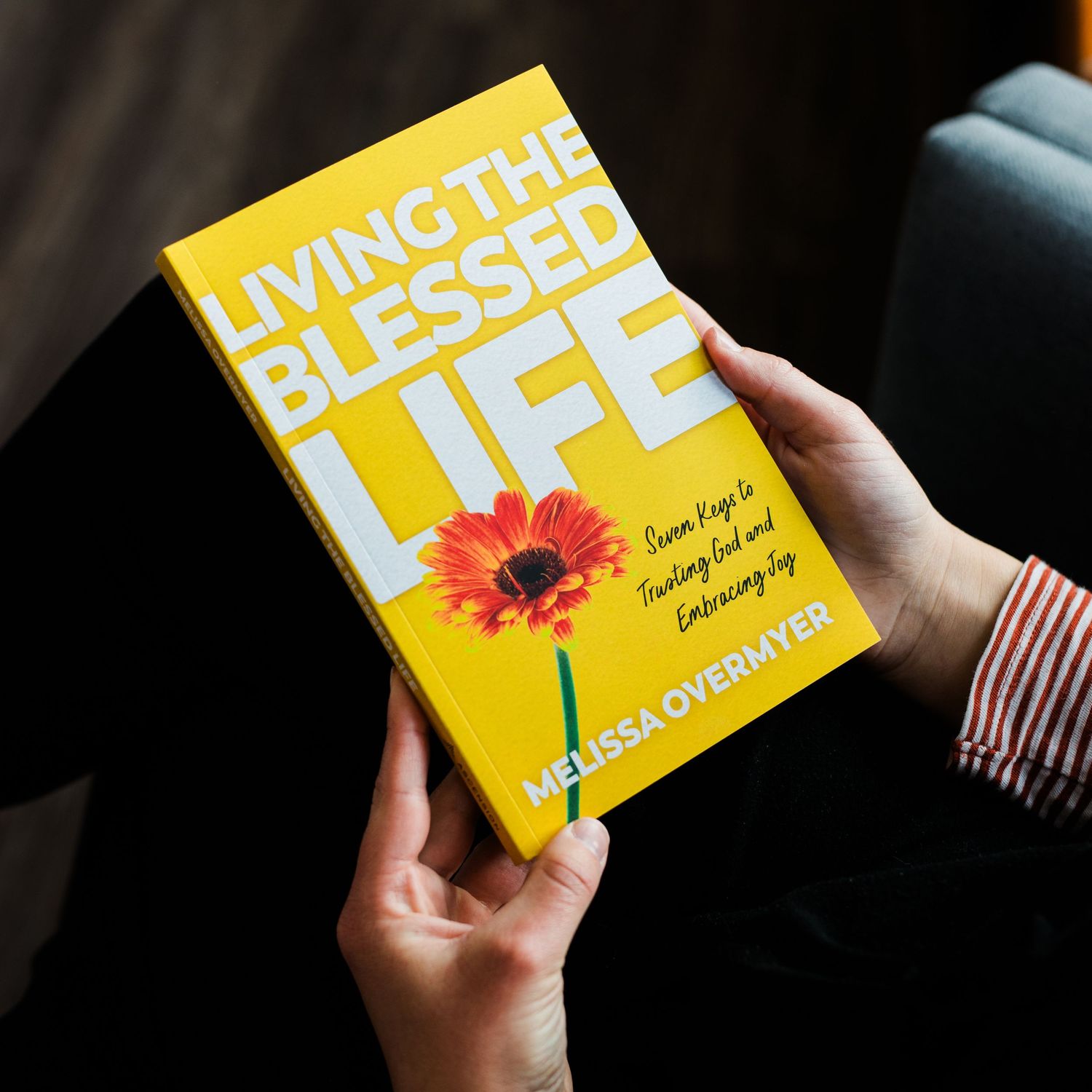 Hands holding the book Living the Blessed Life by Melissa Overmyer with a yellow cover featuring a red flower.