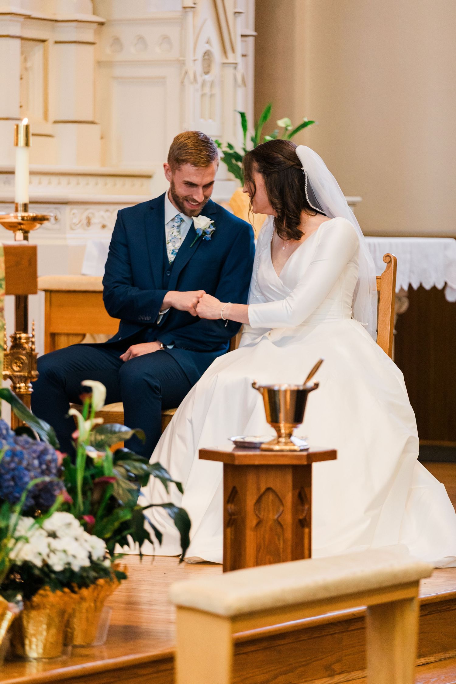 Bride and groom share intimate moment seated at altar during church wedding ceremony with floral arrangements.