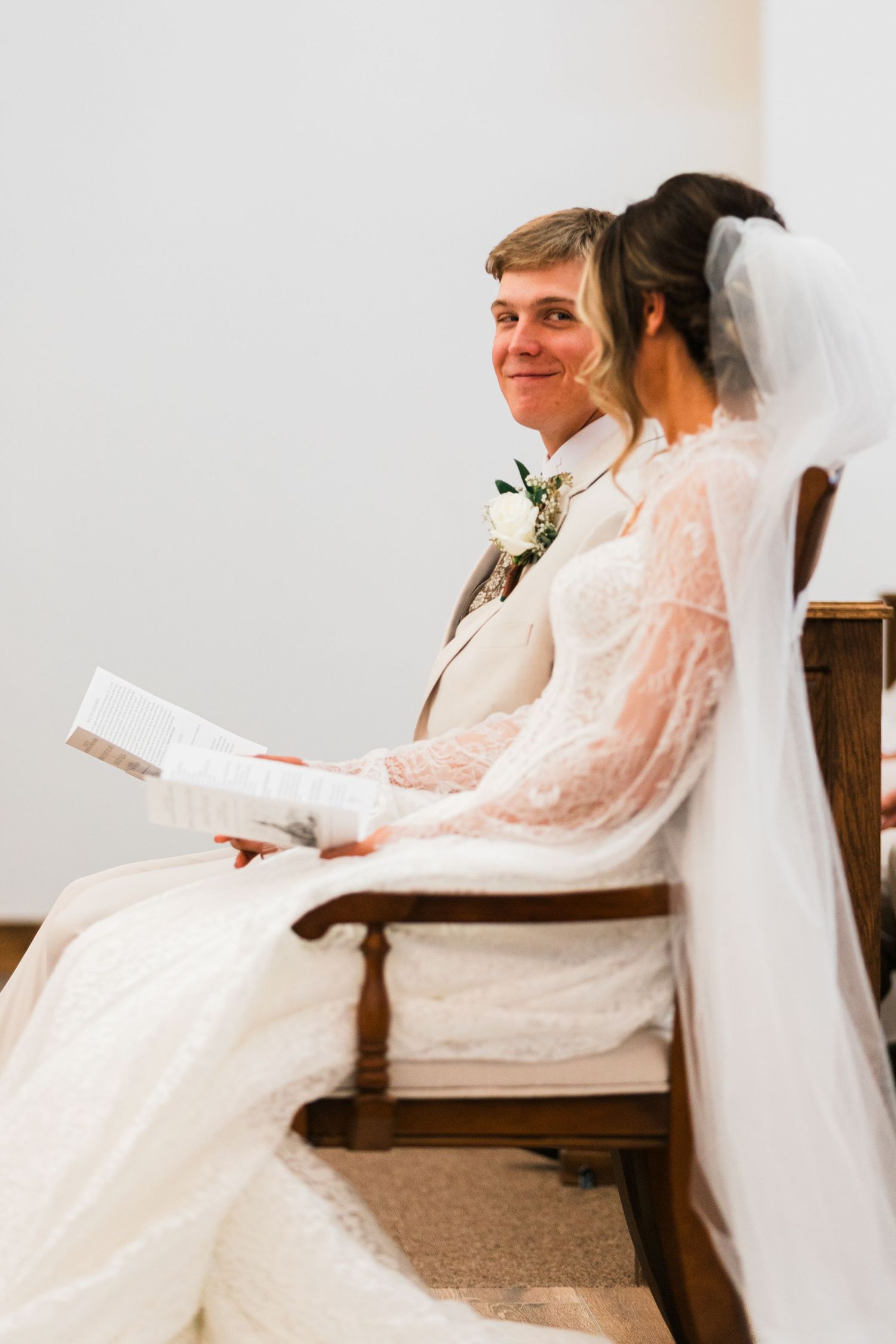 Bride and groom sitting together in church pew during wedding ceremony, groom smiling in suit with boutonniere.