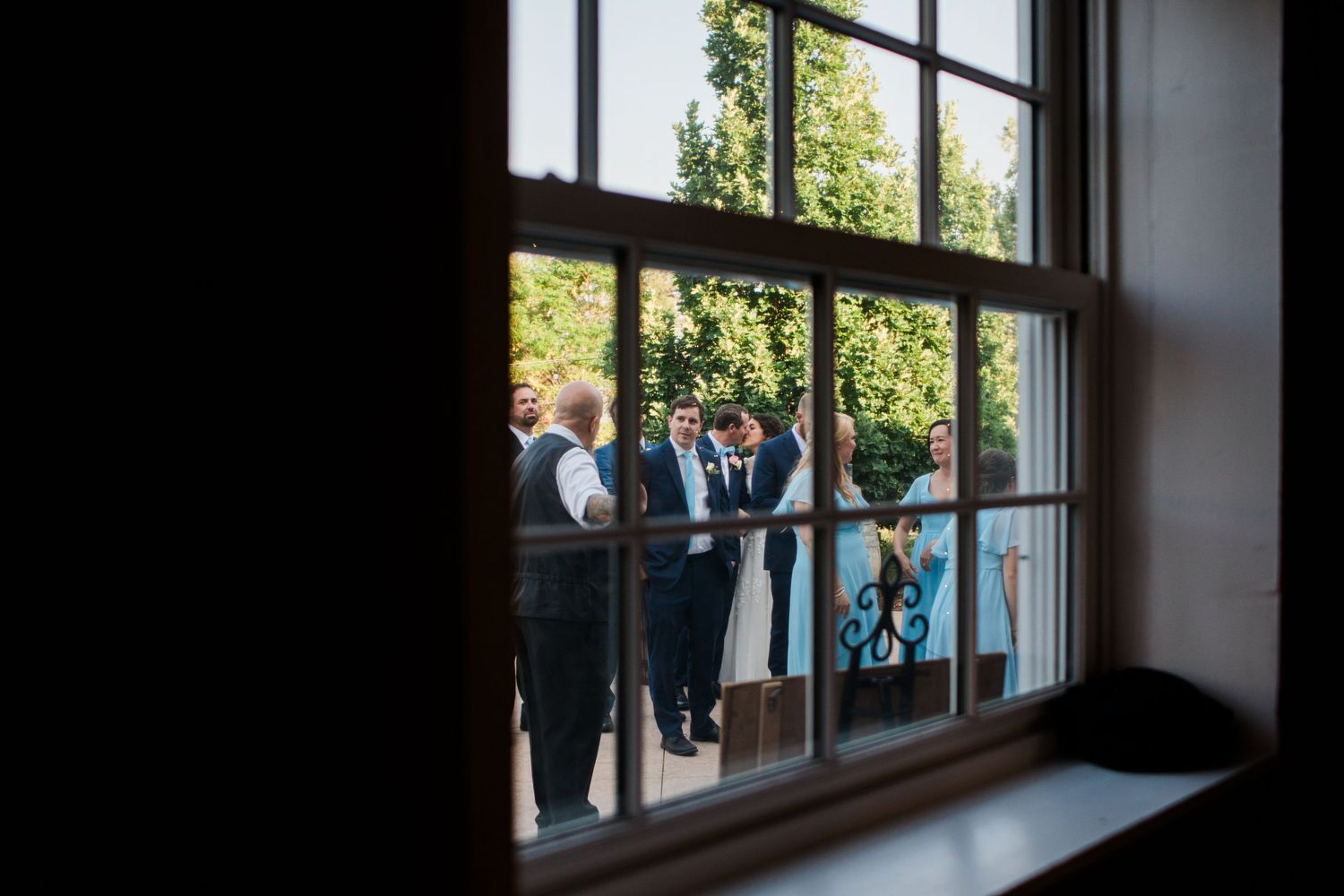 Wedding guests socializing outdoors viewed through a classic sash window, with lush green trees in the background.