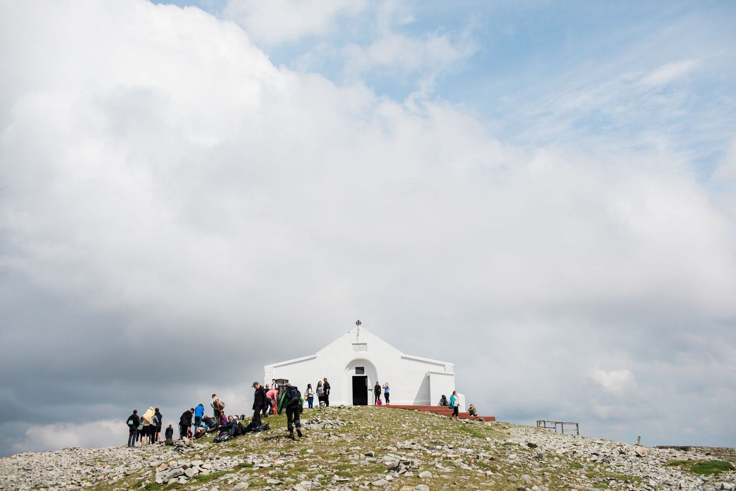 A white hilltop chapel surrounded by visitors under a dramatic cloudy sky on a rocky mountain summit.