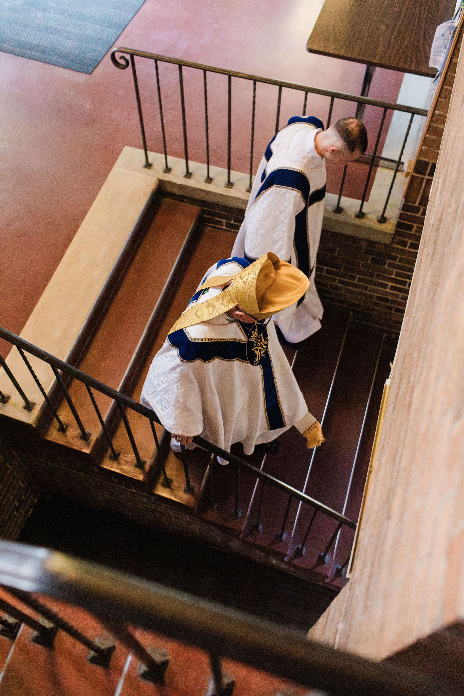 Aerial view of Catholic clergy in white and blue vestments descending ornate brick staircase.