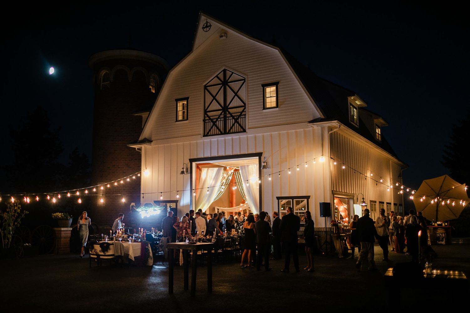 Elegant barn wedding venue glowing with string lights at night, guests mingling outside under a full moon.