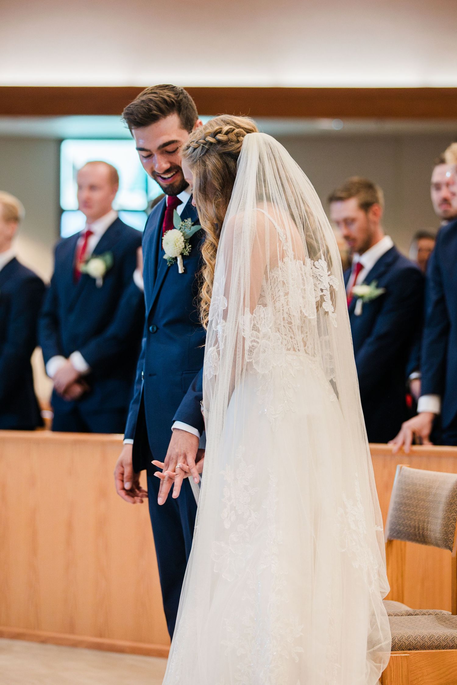 Bride and groom exchange rings at church altar, groomsmen in navy suits visible in background.