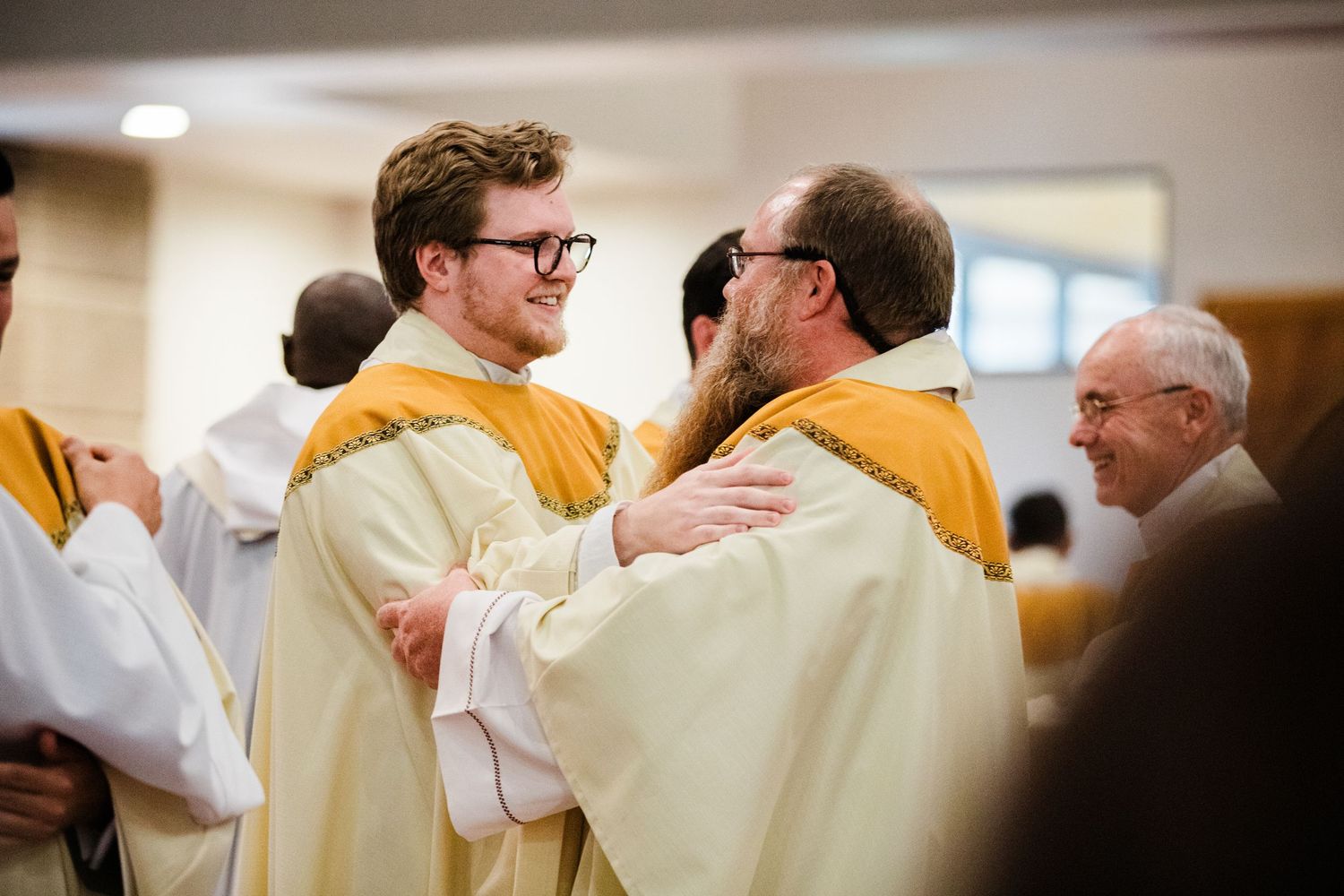 Two men in white and gold vestments share a warm embrace during a Catholic ordination ceremony.