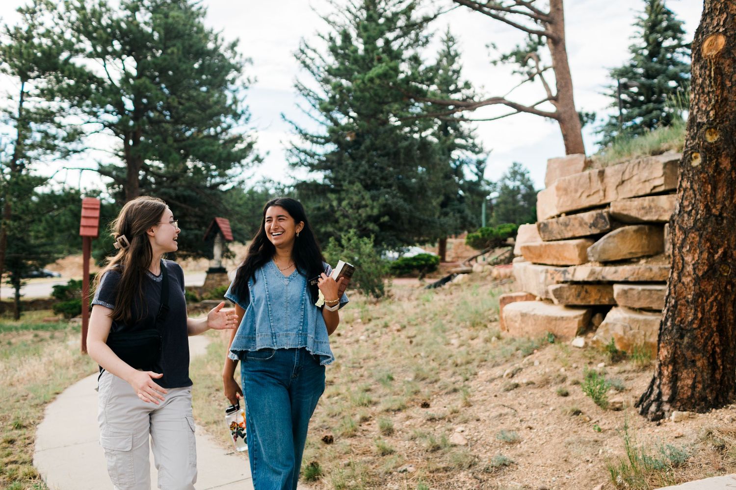 Two women walking and talking on a sunny path surrounded by pine trees and rocky landscape.