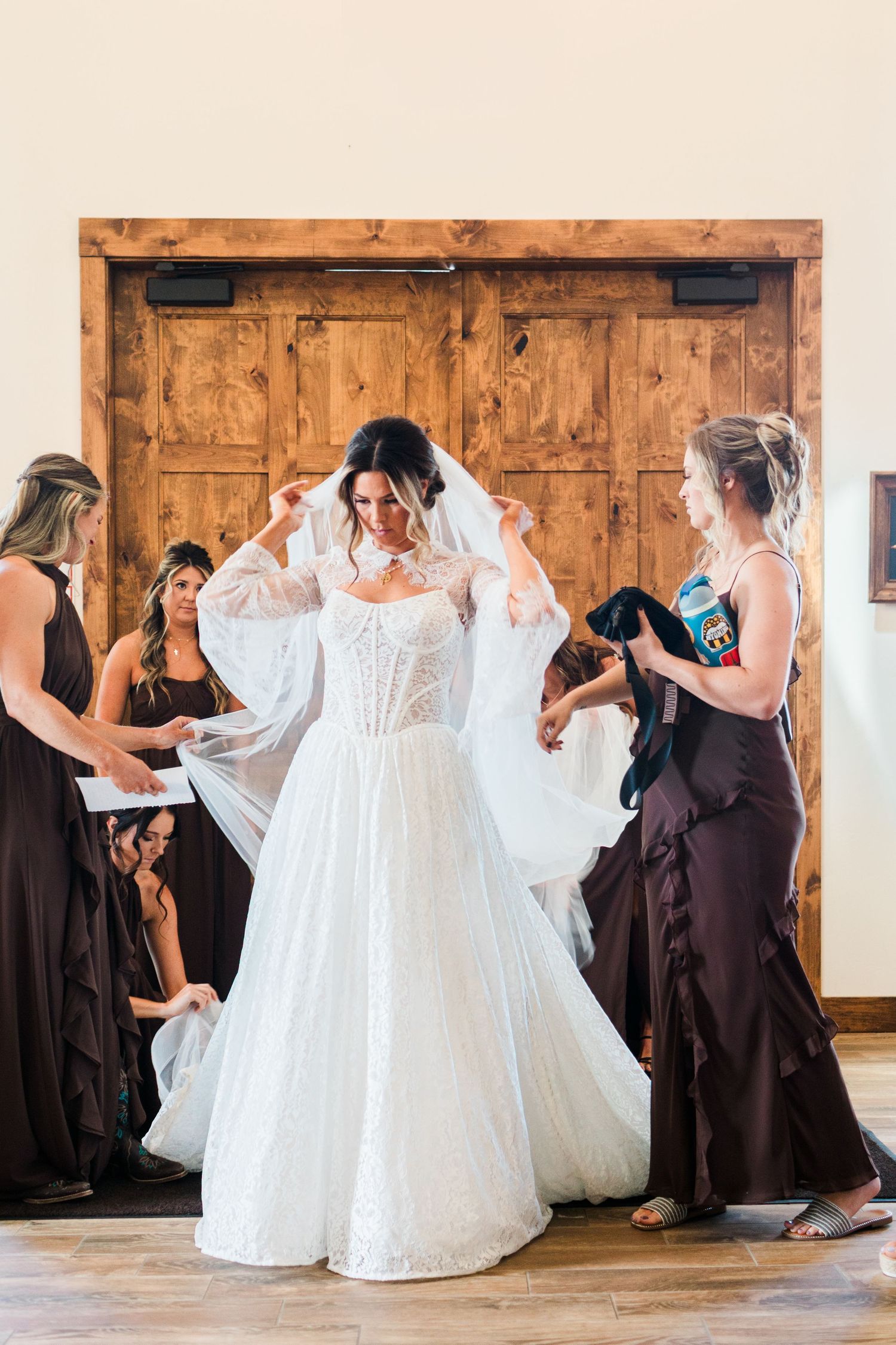 Bride in white lace gown with veil being adjusted by bridesmaids in dark dresses before wedding ceremony.